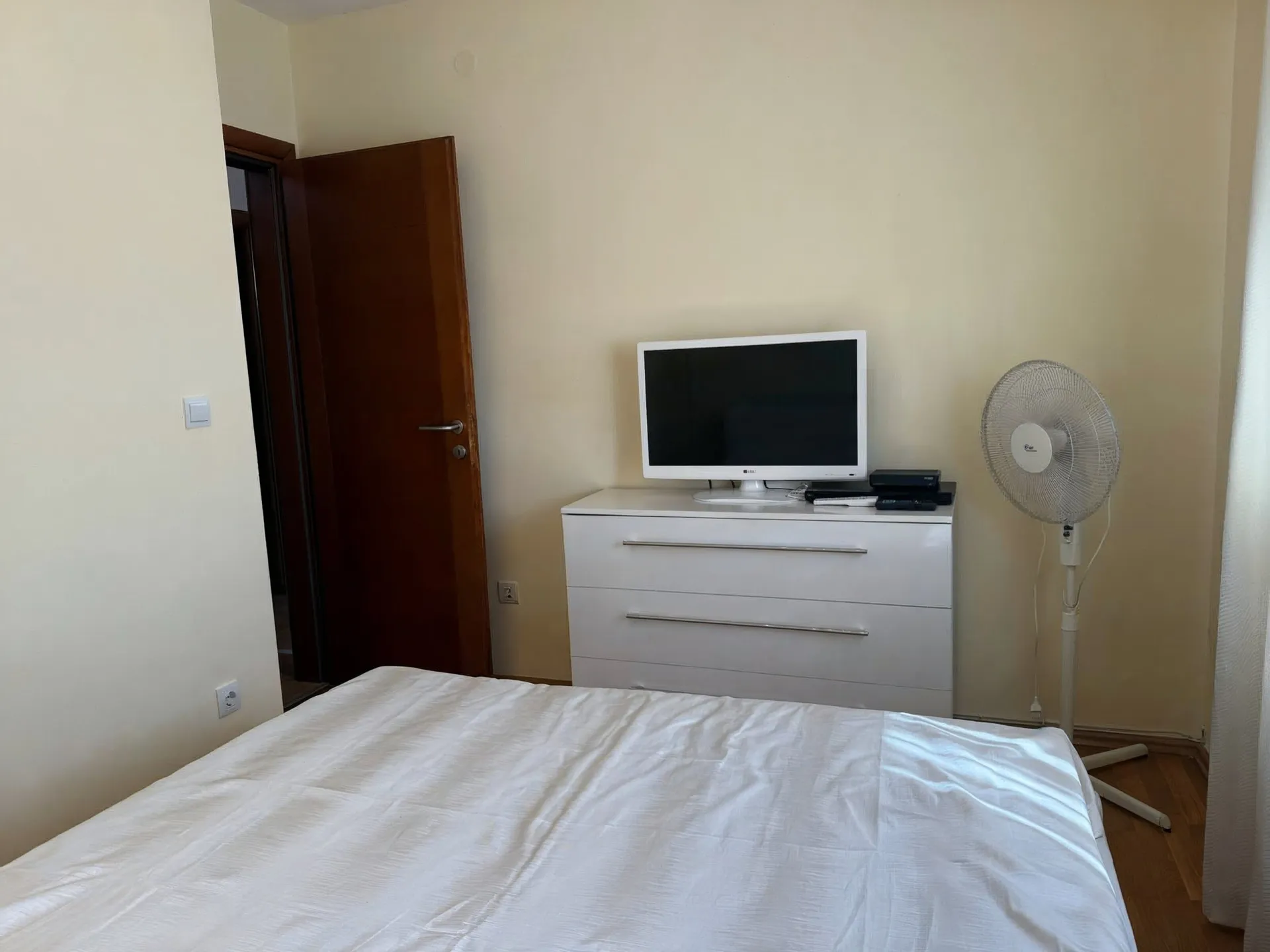 Bedroom with a white bed, a dresser holding a TV, and a standing fan near a light wall.