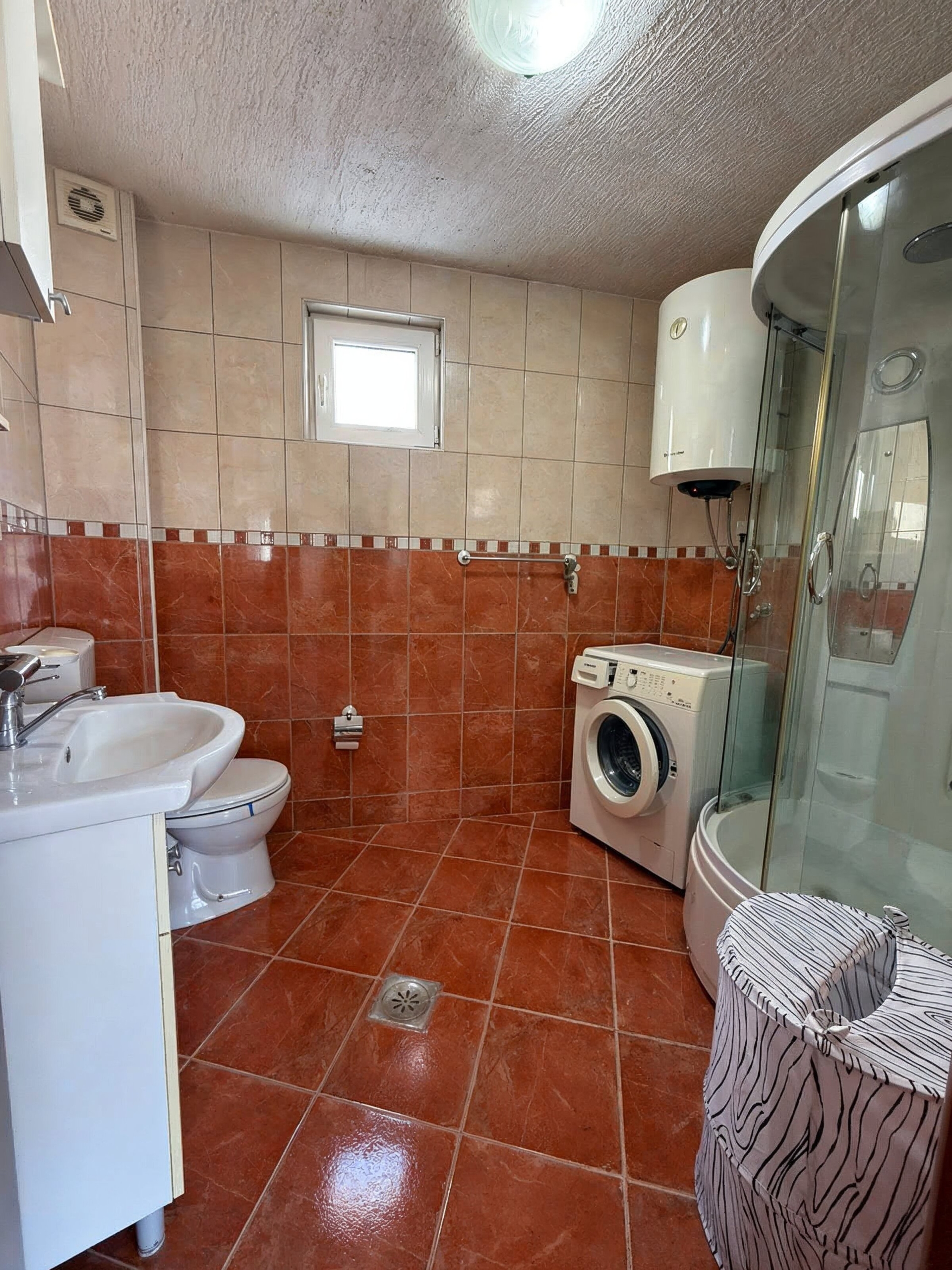 Badezimmer in einer Wohnung in Bečići, Montenegro: Duschkabine, Waschmaschine, Boiler und Fliesen in Terrakottatönen.