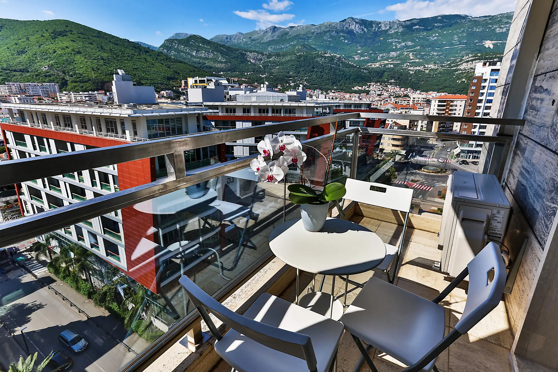 Balkon mit Möbeln und Blick auf die Berge und die Stadt Budva, Montenegro. Auf der Terrasse ein Tischchen mit einer Orchidee und moderne Stühle.