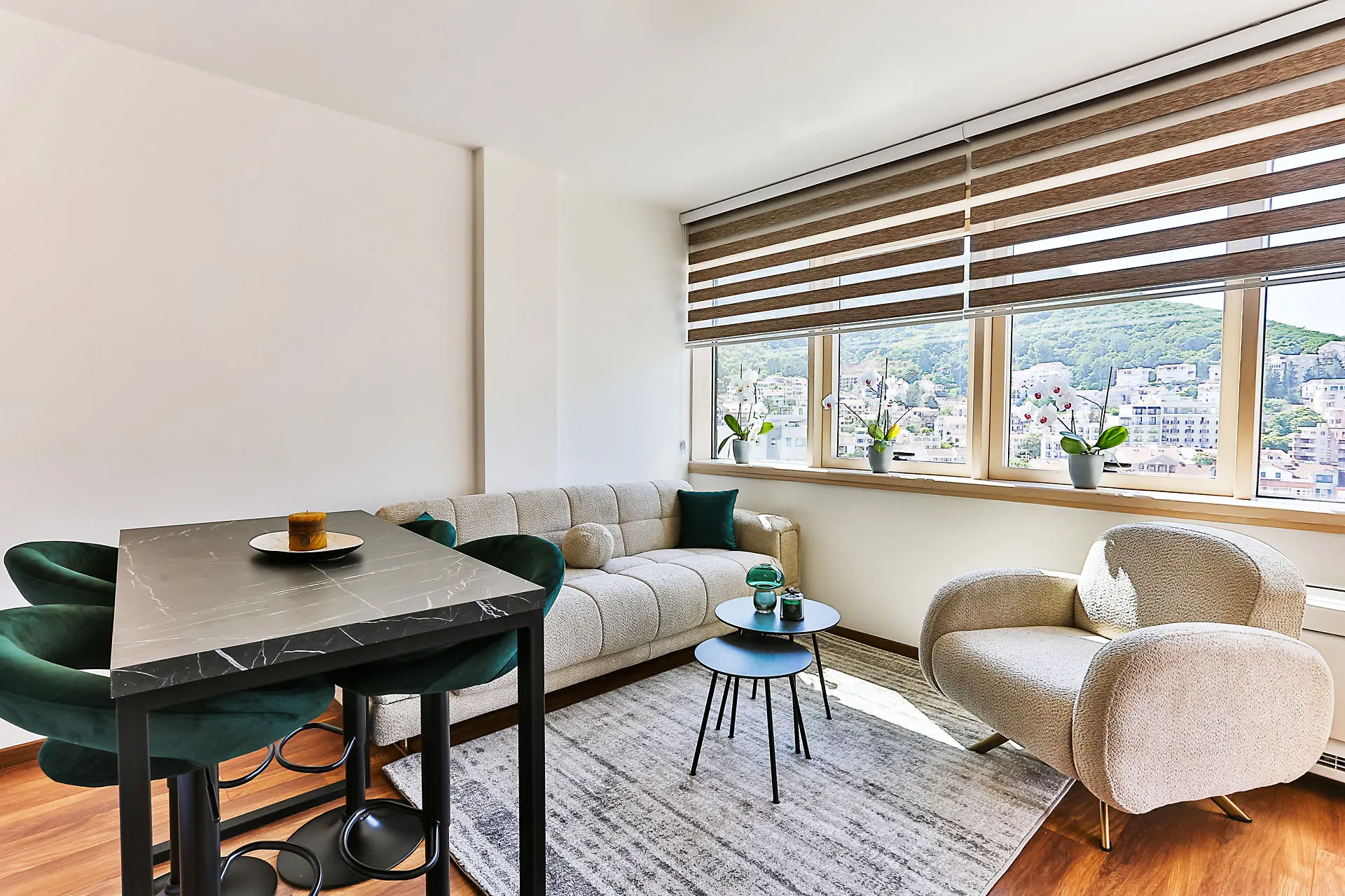 Modernes Wohnzimmer in einer Wohnung in Budva, Montenegro: Essbereich, helles Sofa, Sessel und Bergblick aus dem Fenster.
