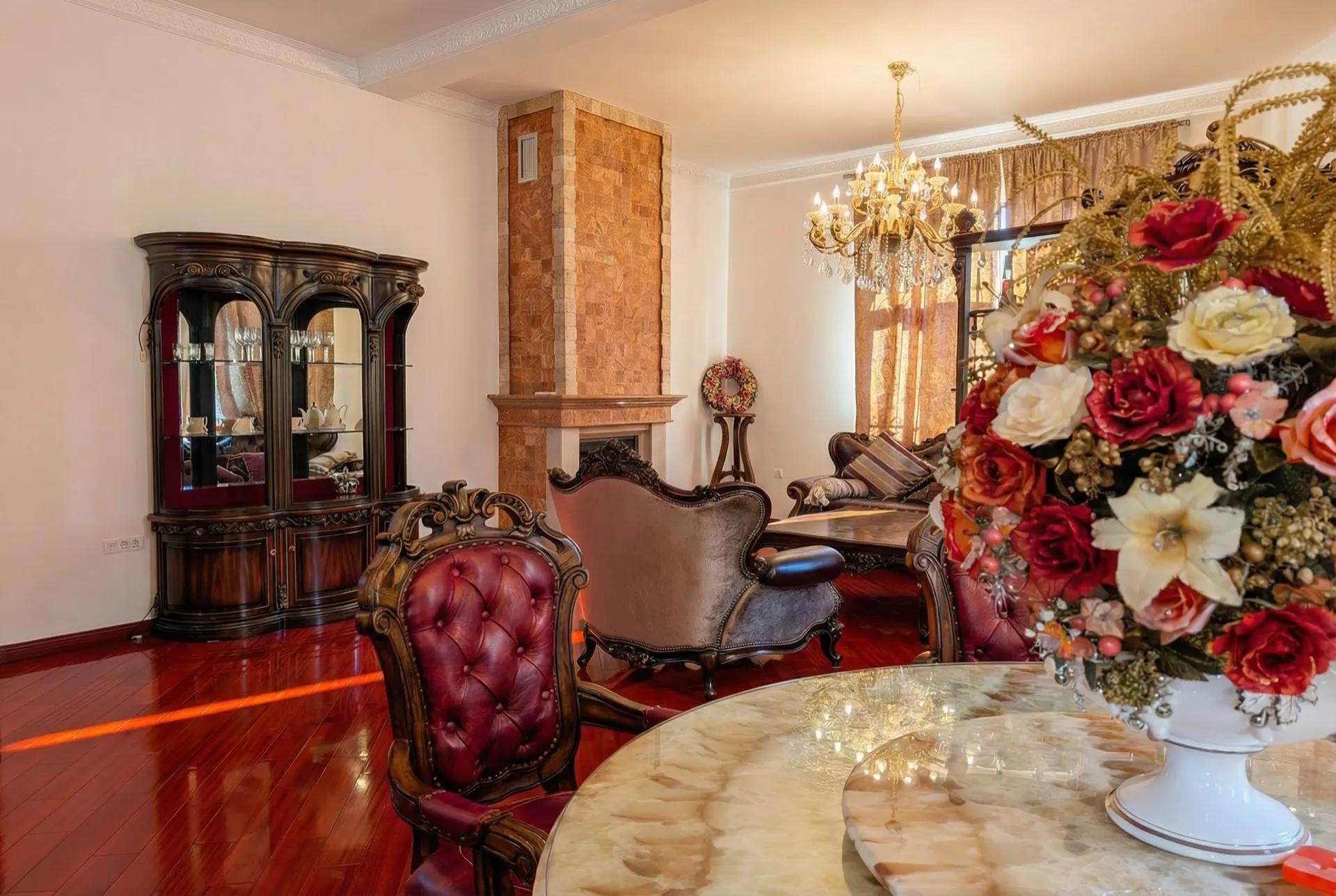 Luxuriöses klassisches Wohnzimmer mit steinerner Kaminwand, Vitrinenschrank, roten Ledersesseln, Kristalllüster und poliertem Parkettboden.