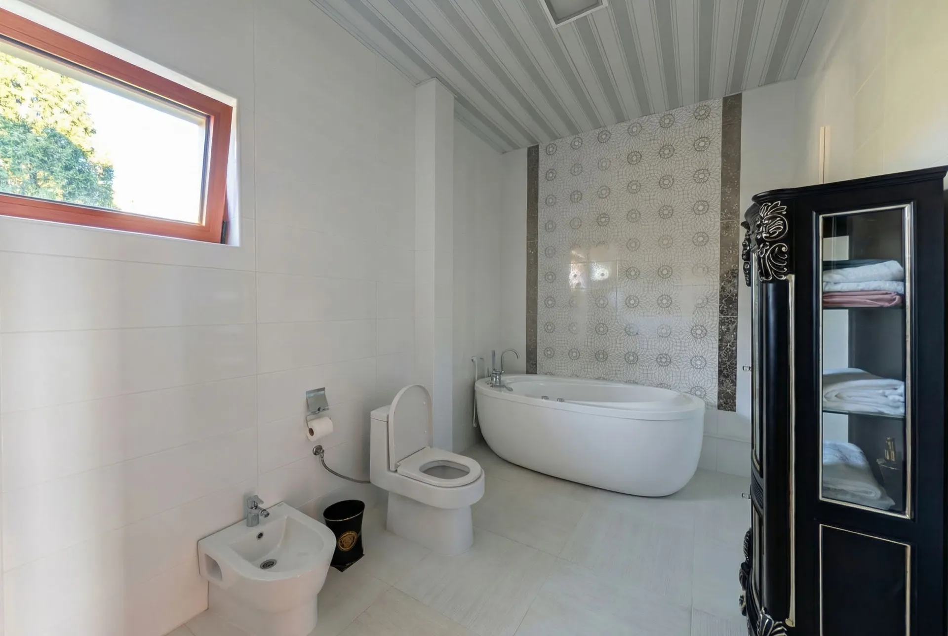 Helle Badezimmer mit weißem Freistehbad, Toilette, Bidet und dunklem Handtuchschrank neben einem Fenster.