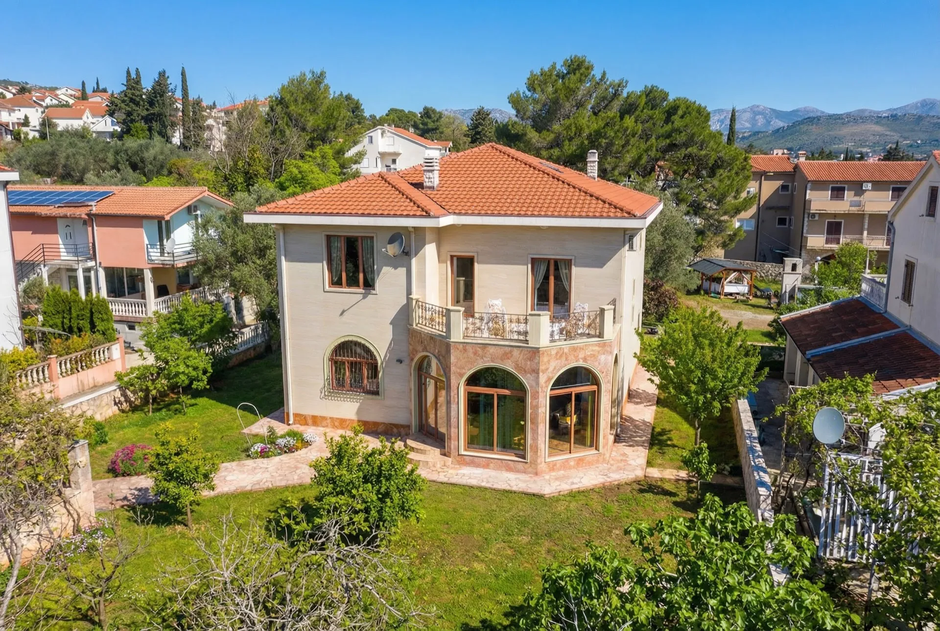 Luftaufnahme eines zweistöckigen Hauses mit rotem Ziegeldach und Balkon, umgeben von einem grünen Garten und anderen Häusern vor Bergen.