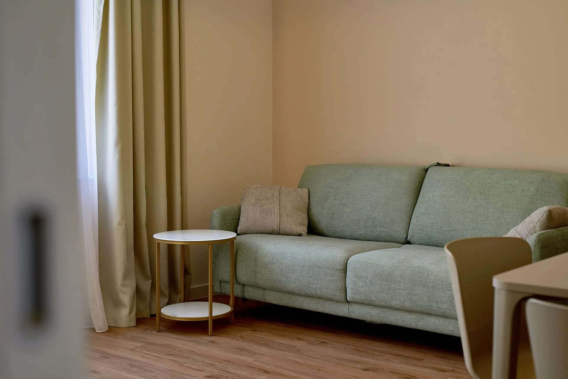 Un canapé vert avec un coussin beige est placé près d'une fenêtre aux rideaux clairs. Une petite table ronde aux pieds dorés se trouve sur le sol en bois.