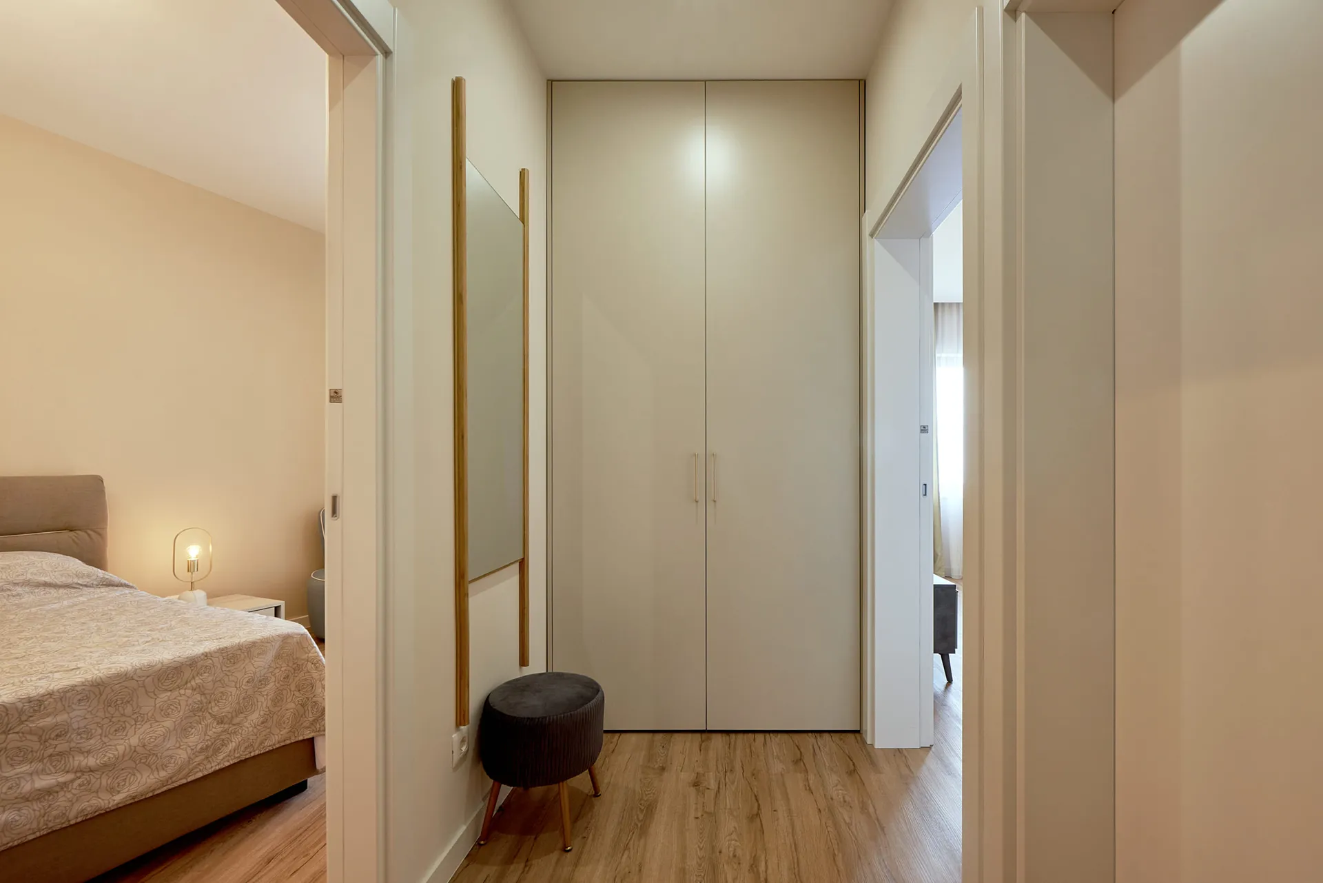 Couloir avec armoire blanche et miroir, vue partielle d'une chambre à coucher avec lit et lampe sur parquet.