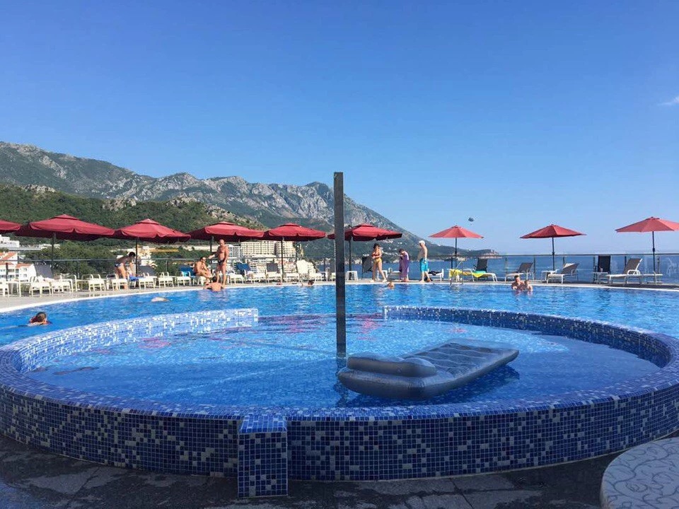 Piscine extérieure avec vue sur la mer et les montagnes dans un complexe résidentiel à Bečići, Monténégro, avec transats et parasols rouges.