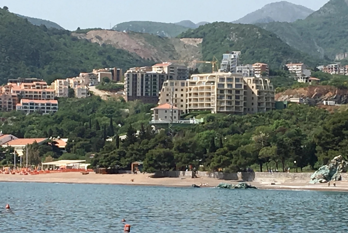 Vue sur le complexe résidentiel et la plage de sable à Bečići, Monténégro : appartements modernes en bord de mer avec les montagnes verdoyantes en arrière-plan.