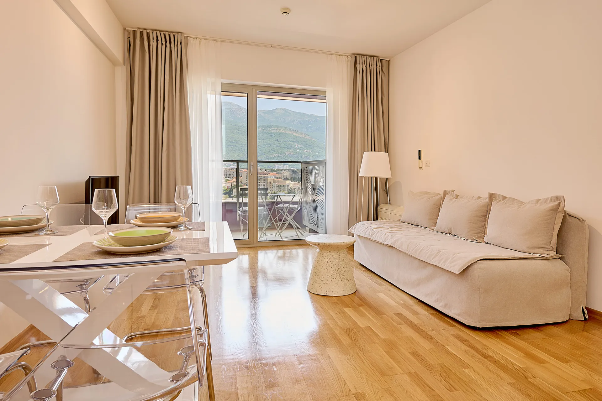 Helle Wohnzimmer mit beigefarbenem Sofa, weißem Tisch und transparenten Stühlen. Blick auf die Berge durch Fenster.