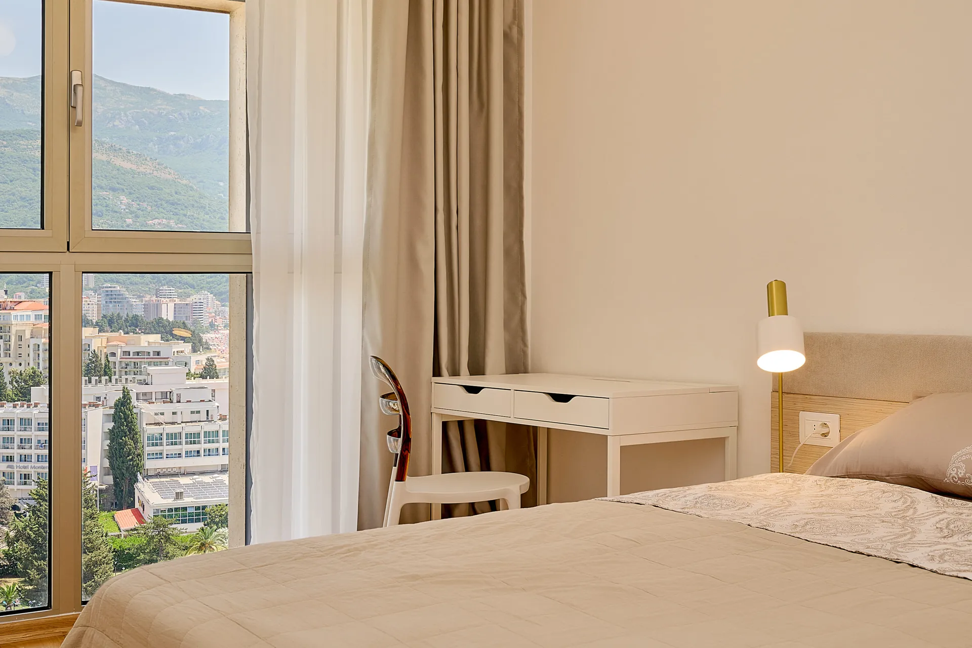 Helles Schlafzimmer mit beigefarbener Bettwäsche, weißem Schreibtisch und Stuhl am Fenster mit Stadt- und Bergblick.