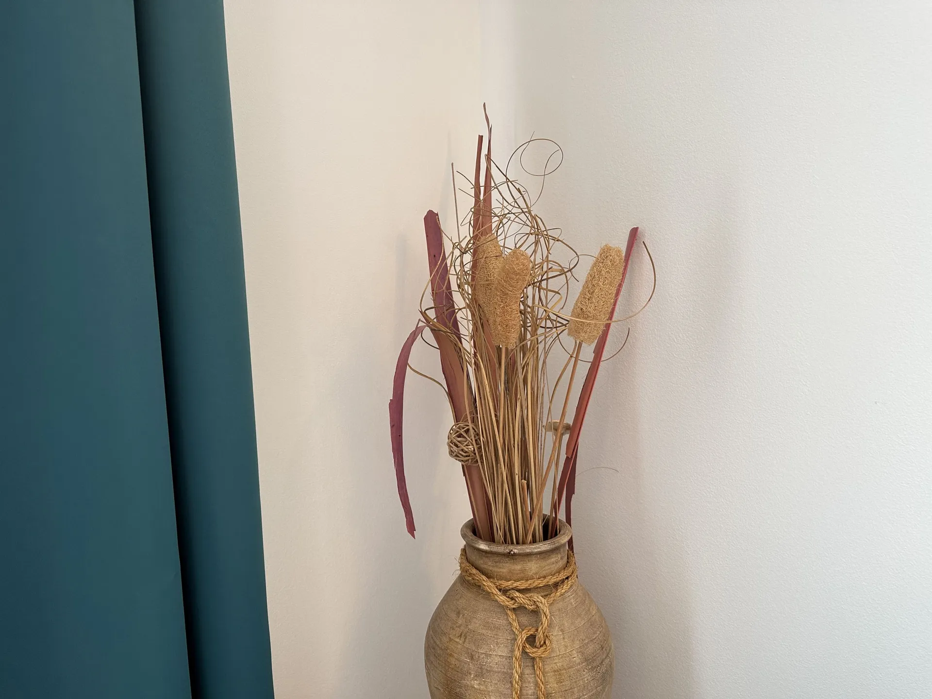 Dekorative Vase mit getrockneten Pflanzen und Seil vor weißer Wand und blauem Vorhang.