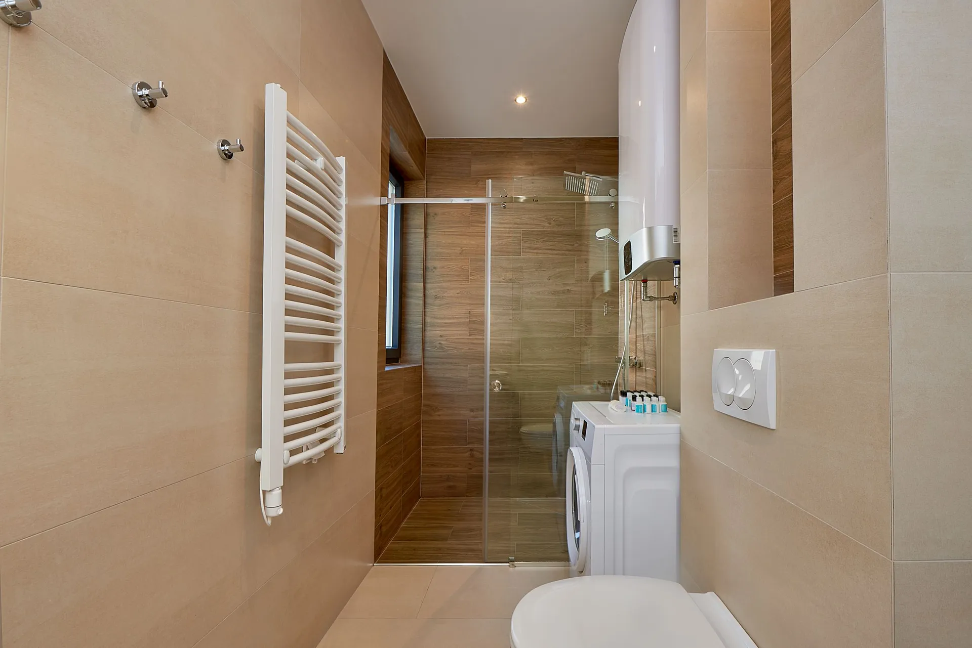 Moderne Baderum mit Glasdusche, Waschmaschine, beheiztem Handtuchhalter und Toilette an beigefarbenen Wänden.