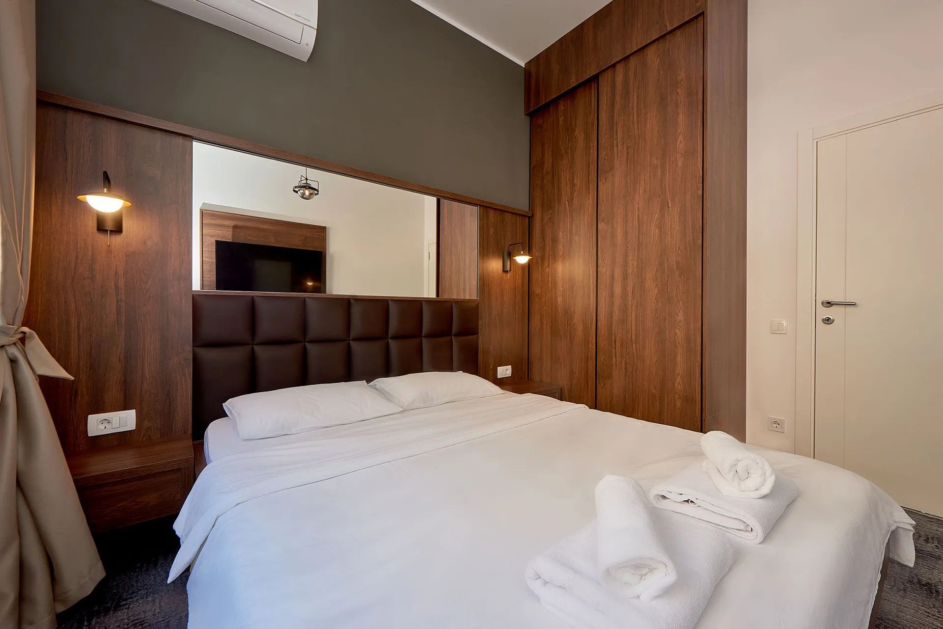 Moderne Schlafzimmer mit Holzverkleidung, großem Spiegel und weißer Bettwäsche. Auf dem Bett liegen gerollte Handtücher.
