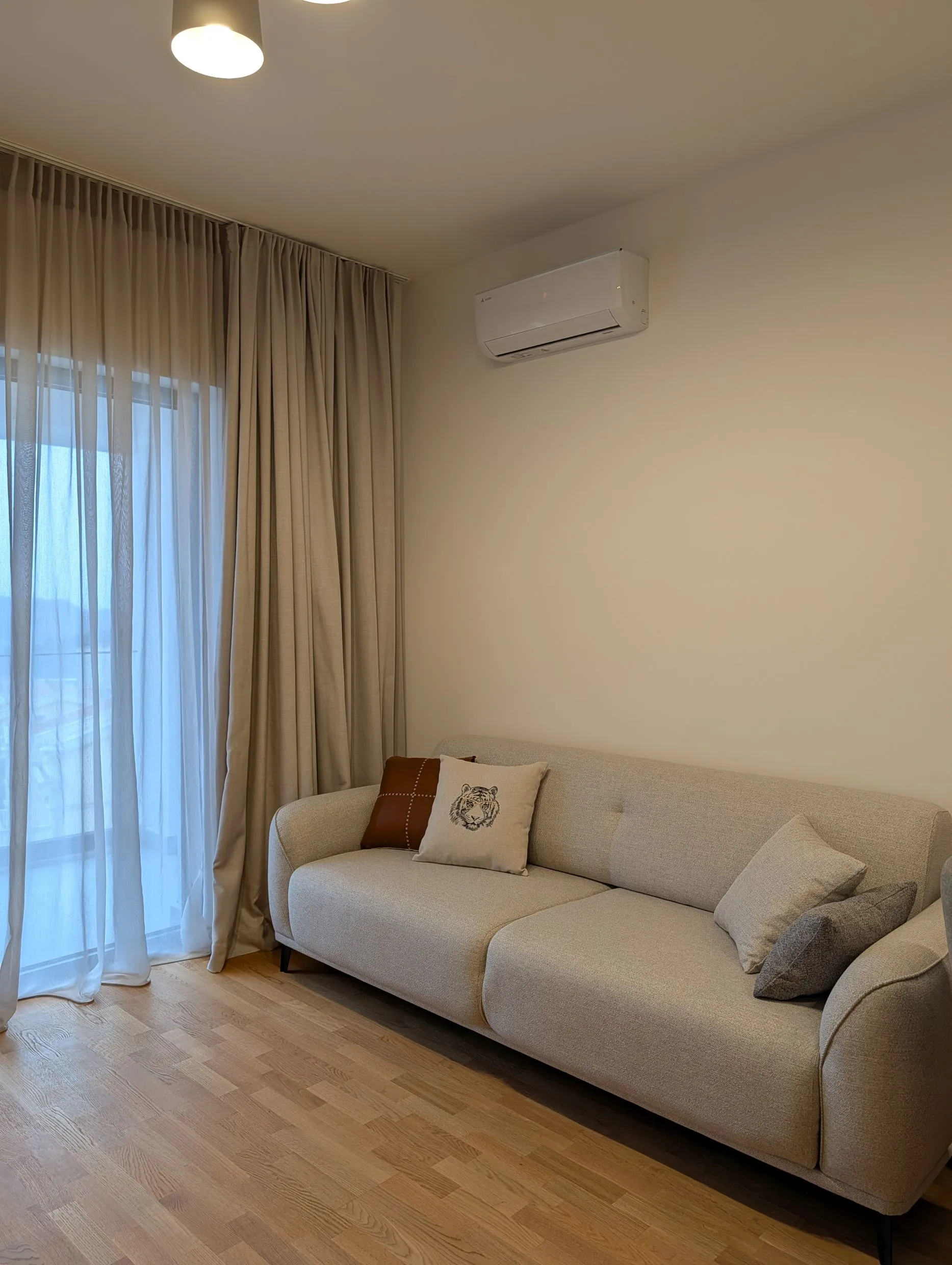 Hellraum mit beigefarbenem Sofa, Tigerkissen, Holzboden und Wand-Klimaanlage.