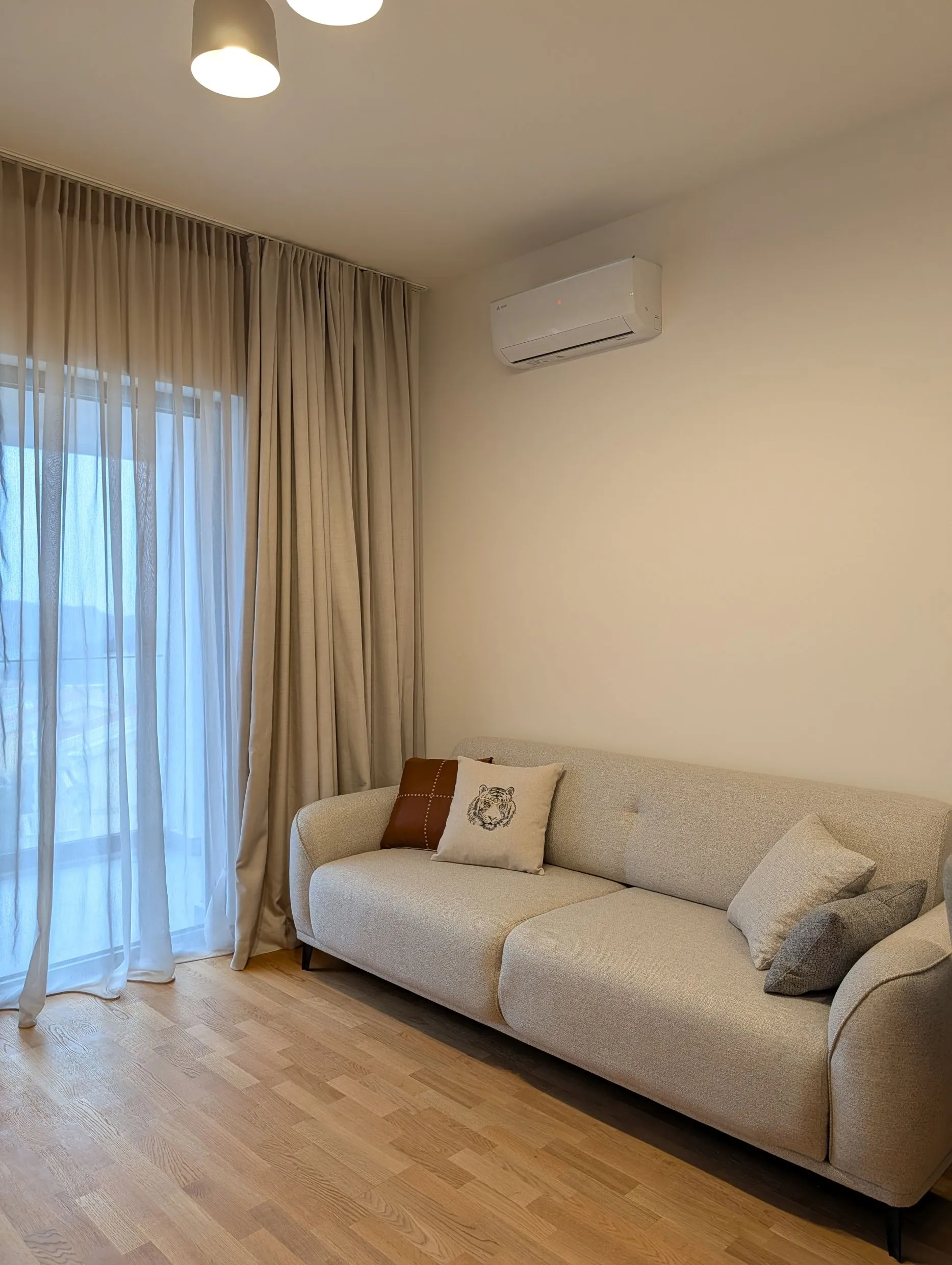Hellraum mit beigem Sofa, Tigerkissen, Holzboden und Wand-Klimaanlage.