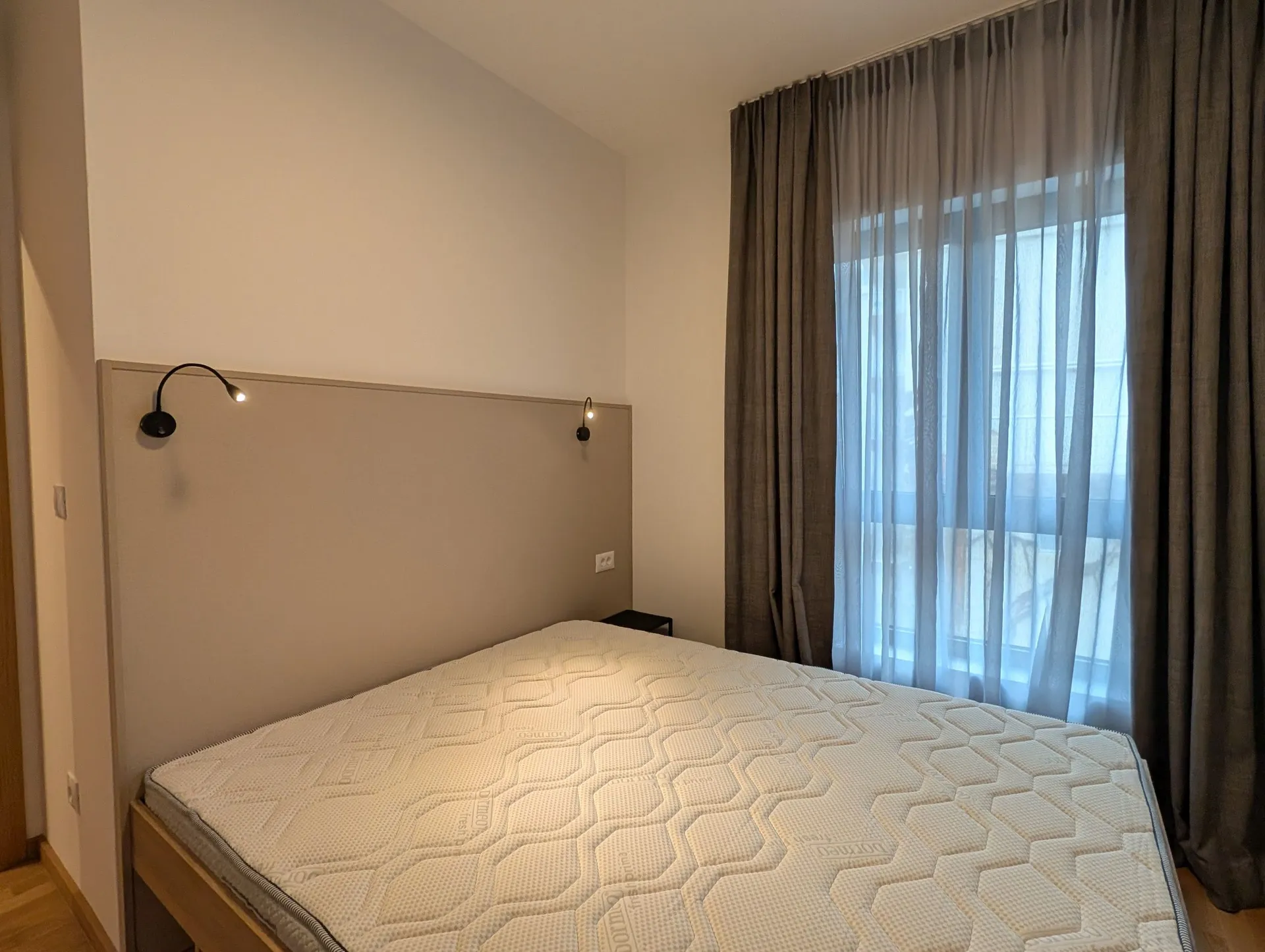 Moderne Schlafzimmer mit weißer Matratze, grauem gepolstertem Kopfteil, Wandleuchten und einem Fenster mit Vorhängen.