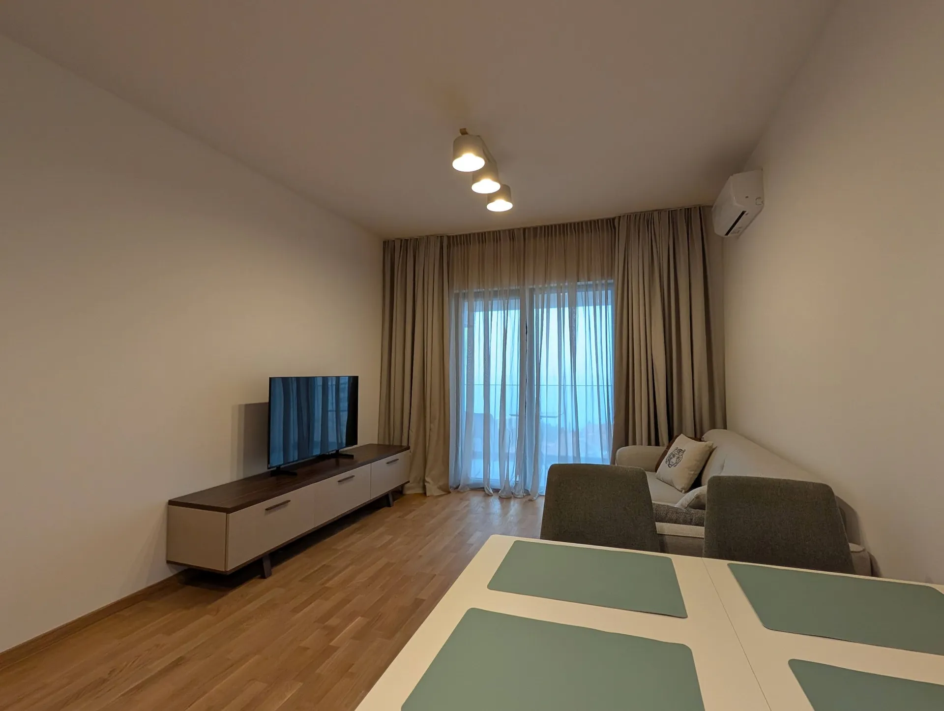 Helle Wohnzimmer mit Sofa, Fernseher auf Konsole und Esstisch neben einem Fenster mit Vorhängen.