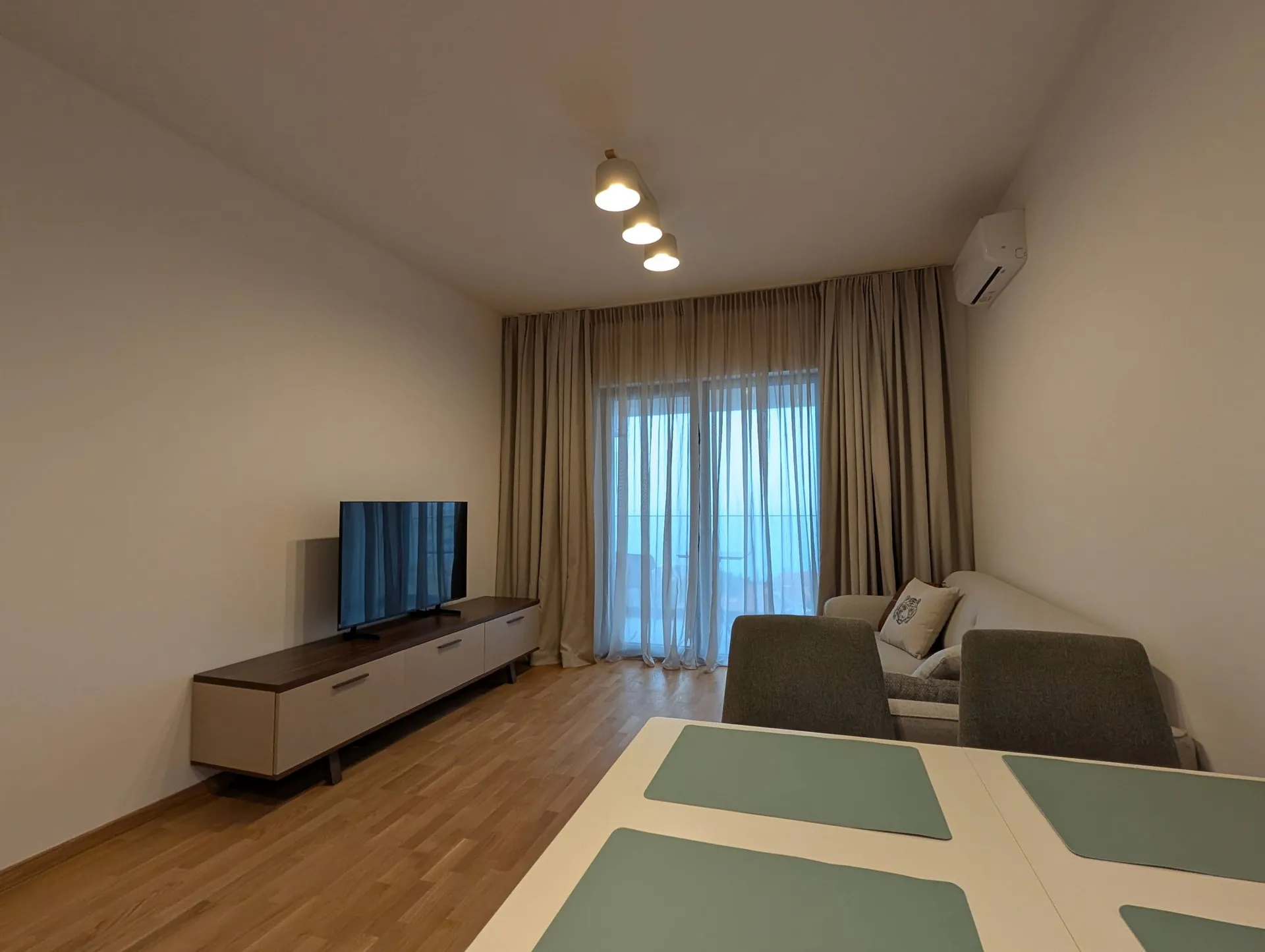 Helles Wohnzimmer mit Fernseher auf Konsole, Sofa, Esstisch und großem Fenster mit beigen Vorhängen.