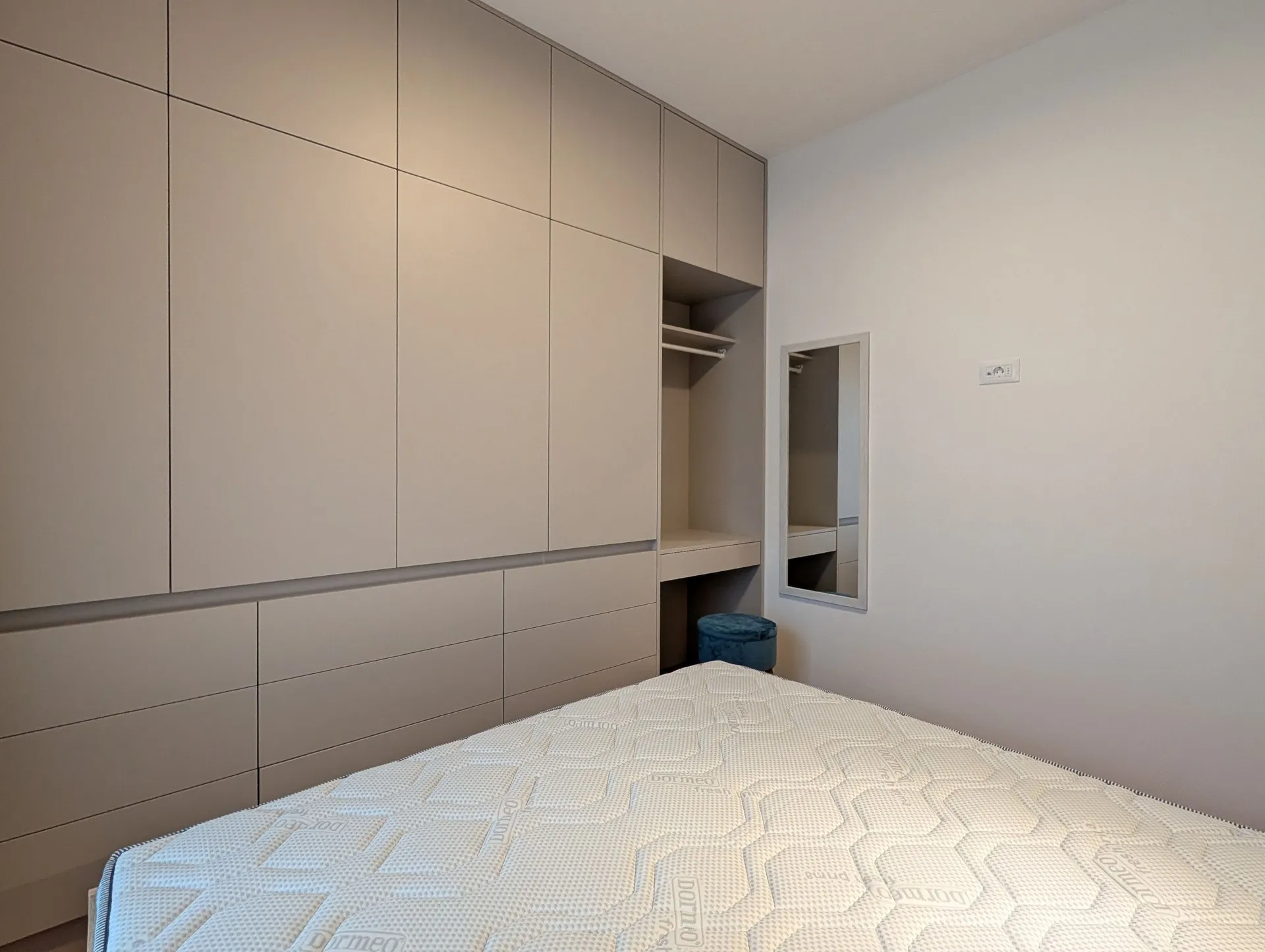 Moderne Schlafzimmer mit beigefarbener Einbaugarderobe, Matratze und kleinem blauen Hocker.