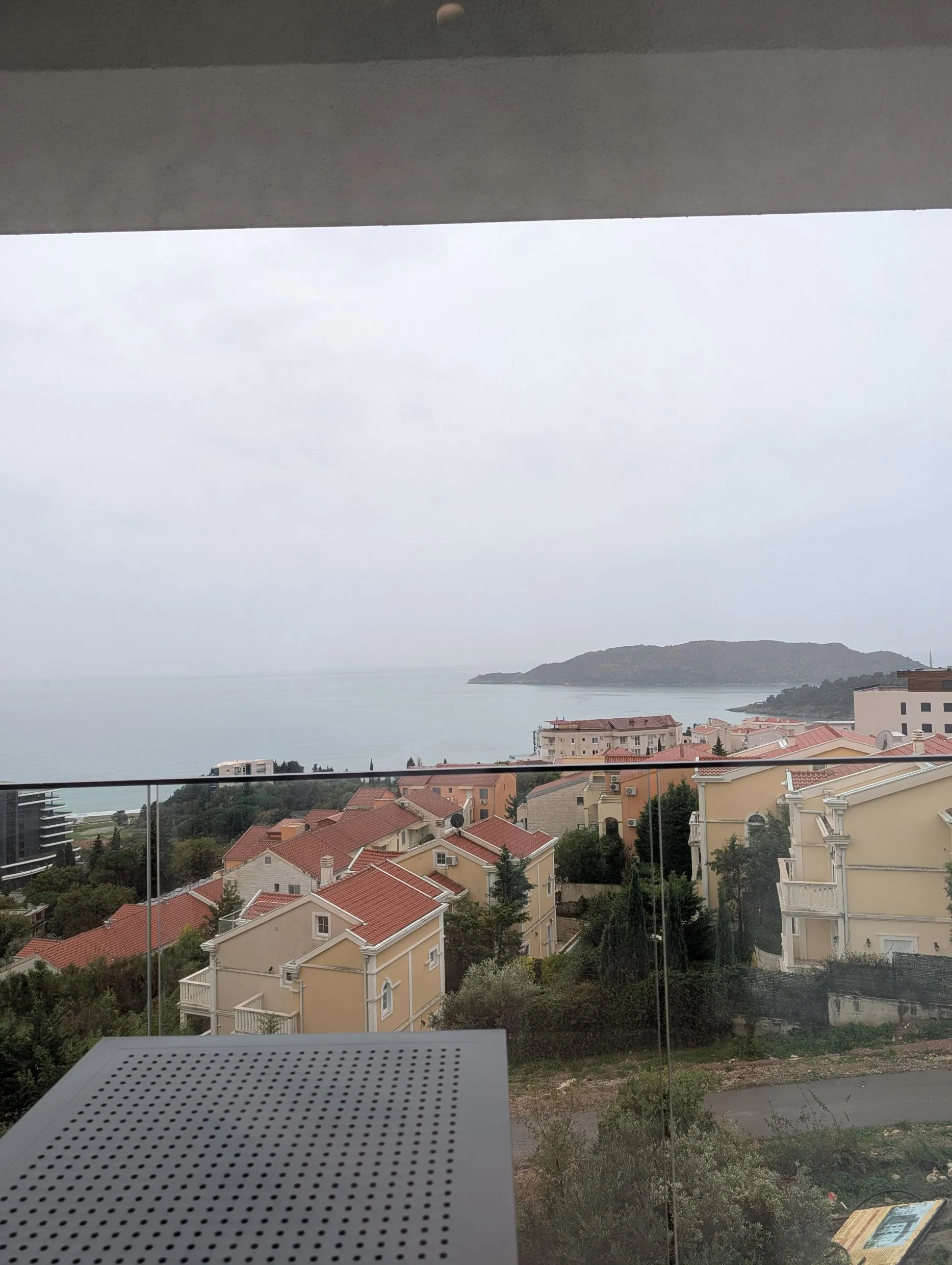 Blick vom Balkon auf Häuser mit roten Dächern und das Meer an einem bewölkten Tag.