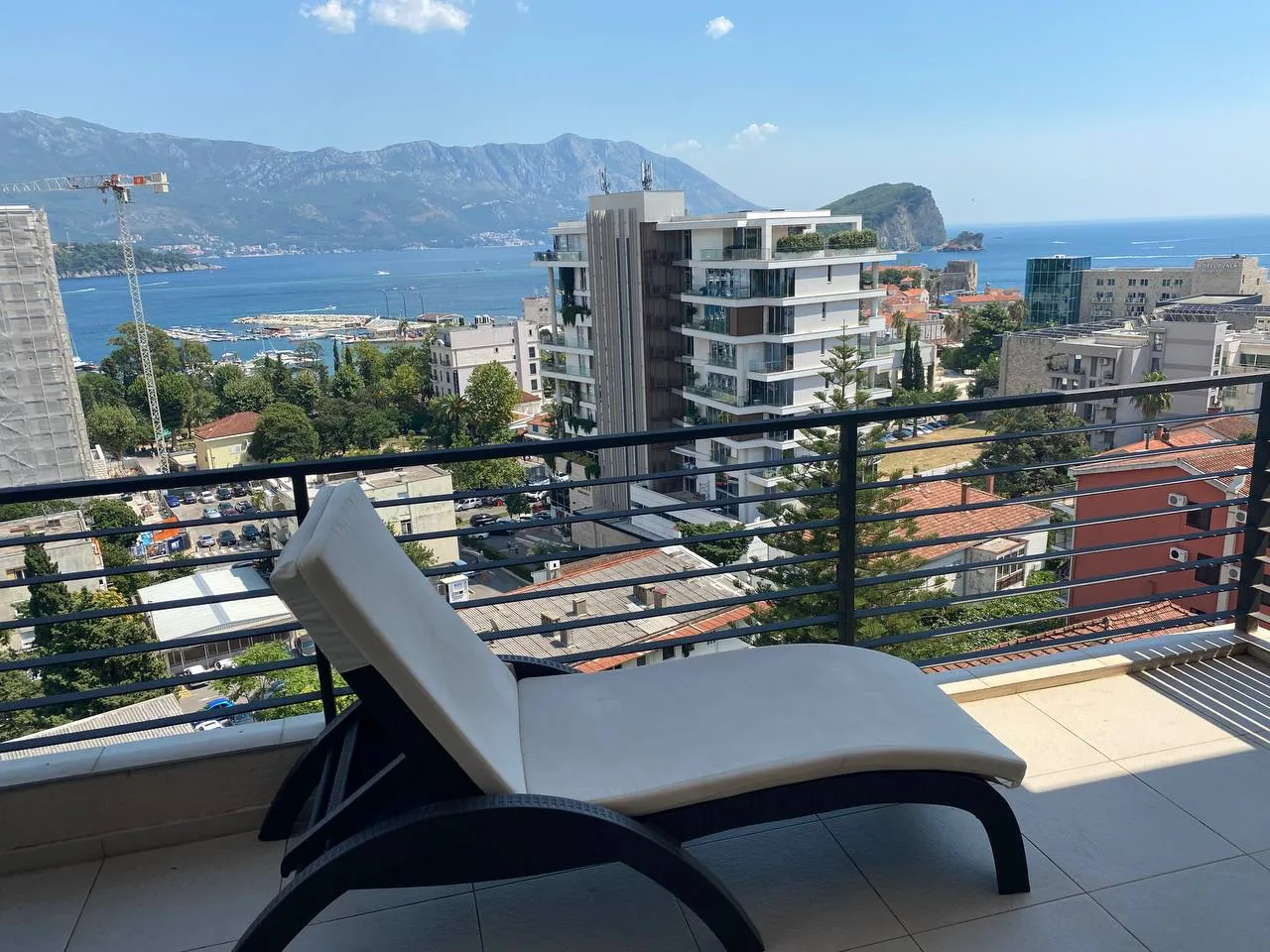 Balcon avec transat offrant une vue sur la mer, les montagnes et les bâtiments urbains.