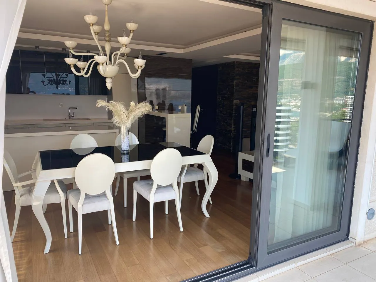 Salle à manger lumineuse avec une table blanche, six chaises et un lustre décoratif. Une cuisine est visible en arrière-plan près d'une grande porte coulissante en verre.