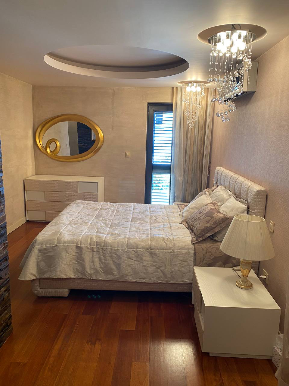 Sypialnia w apartamencie w Budvie, Czarnogóra: łóżko podwójne, szafka nocna, komoda, lustro i kryształowy żyrandol.