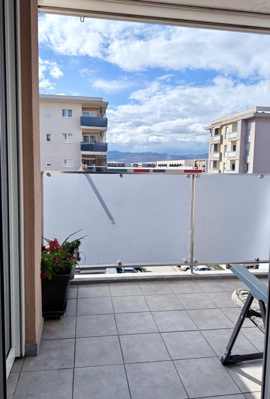 Balcon d'un appartement à Podgorica, Monténégro : sol carrelé, garde-corps en verre et vue sur la ville et les montagnes.