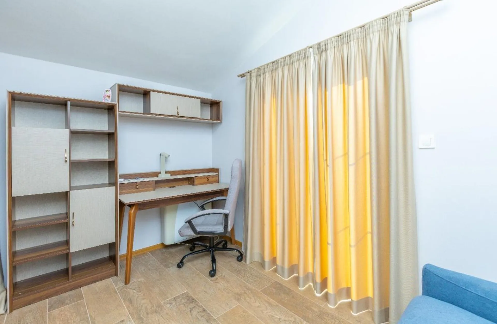 Hellraum mit Holzpult, Bürostuhl, hohem Regal und beigefarbenen Vorhängen vor hellblauen Wänden sowie Fliesenboden.