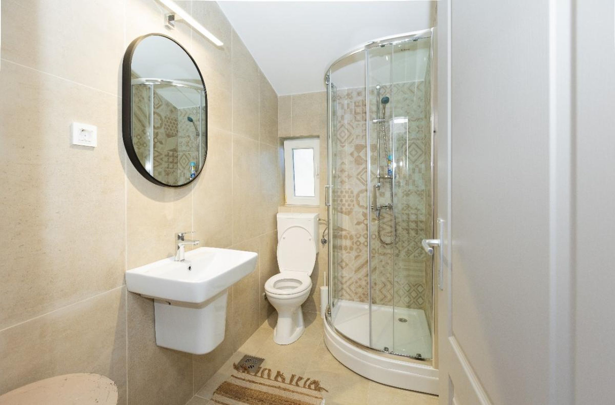 Salle de bains moderne avec cabine de douche dans une maison, ville de Tivat, Monténégro : carrelage beige et miroir rond.