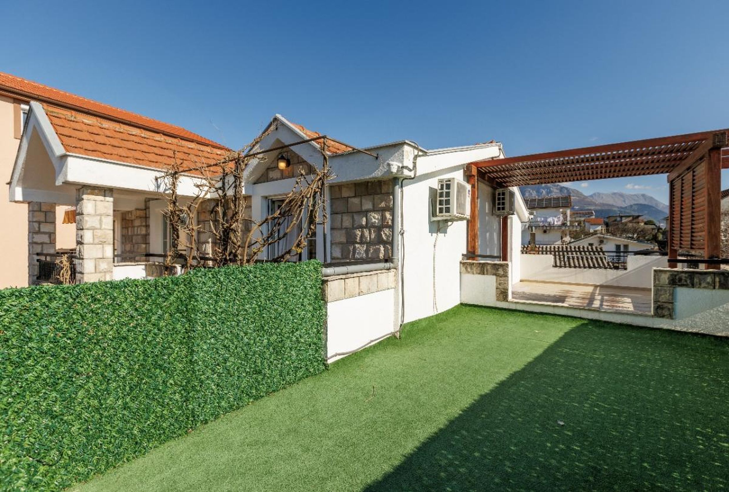 Terrasse de la maison à Tivat, Monténégro : gazon artificiel, pergola en bois, parement en pierre et vue sur les montagnes.