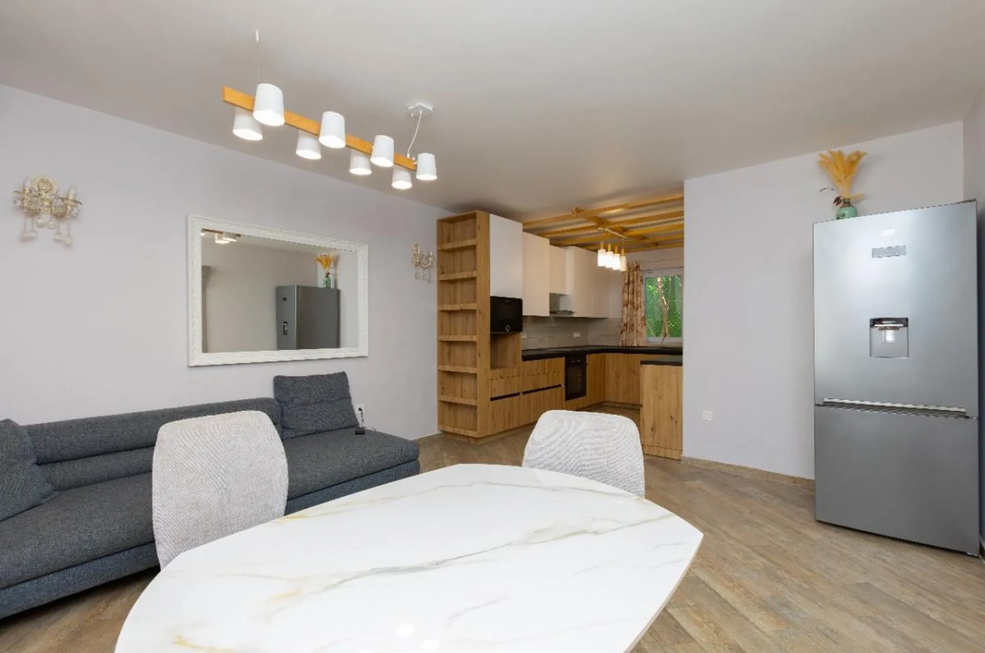 Helle Wohnzimmer mit grauem Sofa, weißem Esstisch und offener Küche mit hellen Holzelementen.