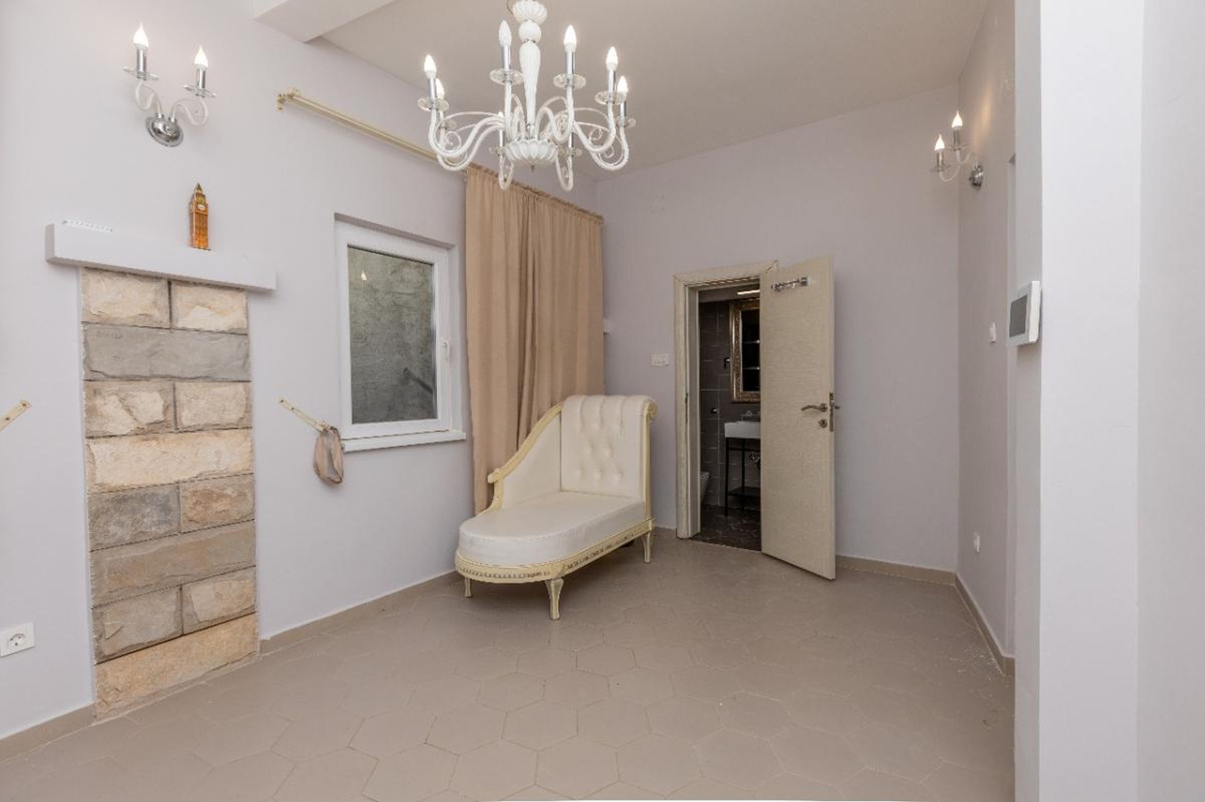 Salon dans une maison à Tivat, Monténégro : canapé classique, mur en pierre, lustre et entrée de la salle de bains.
