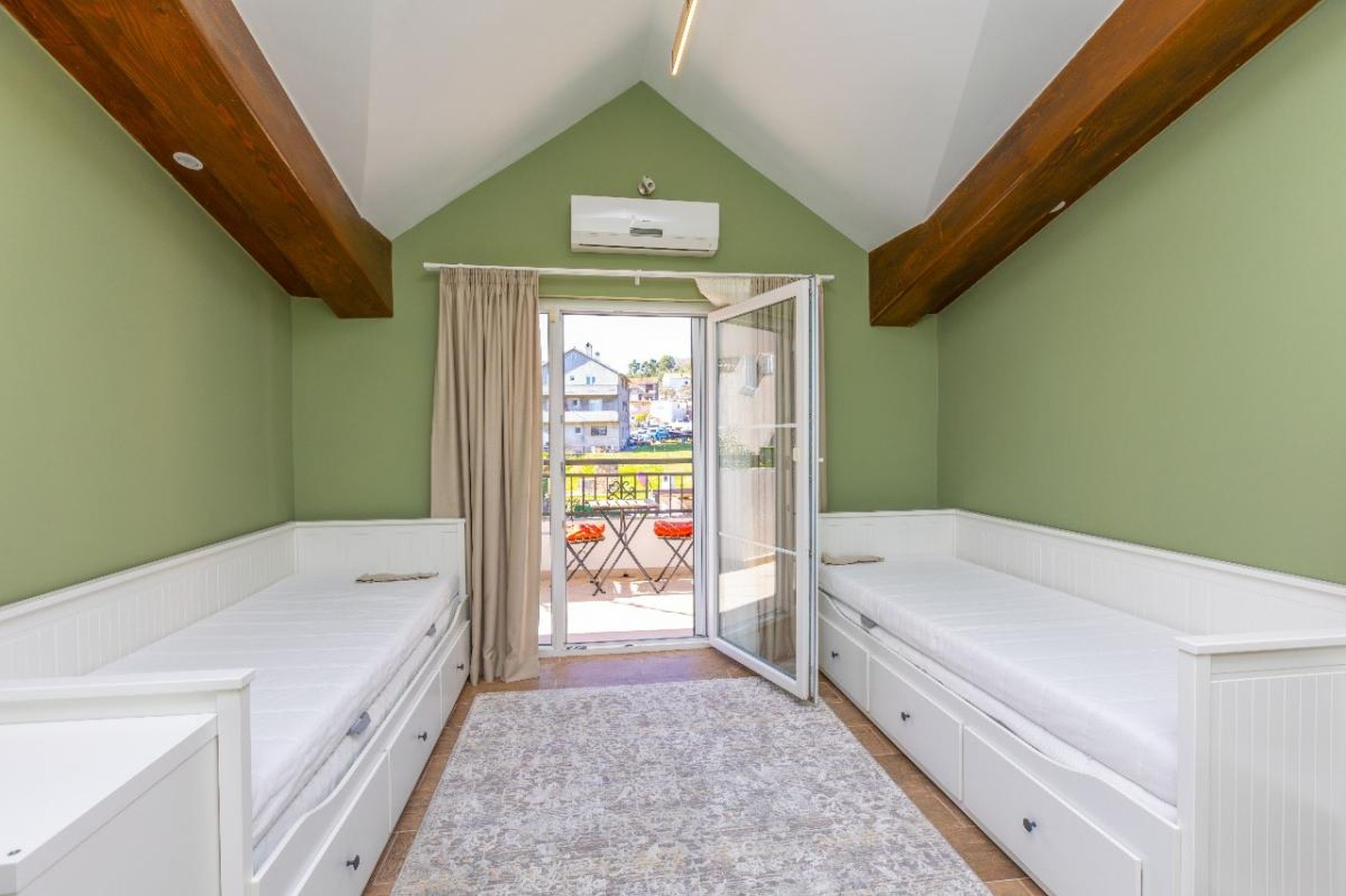 Chambre mansardée dans une maison à Tivat, Monténégro : deux lits blancs, poutres en bois, accès au balcon avec vue.