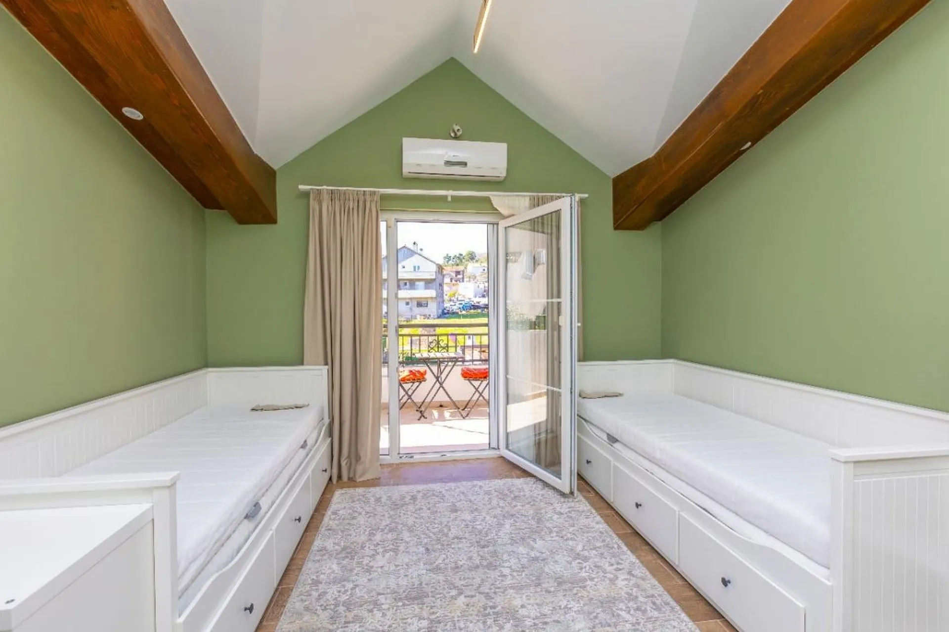 Heller Schlafzimmer mit zwei weißen Betten auf Standschränken, grüne Wände, Holzbalken an der Decke und ein offener Balkonzugang.