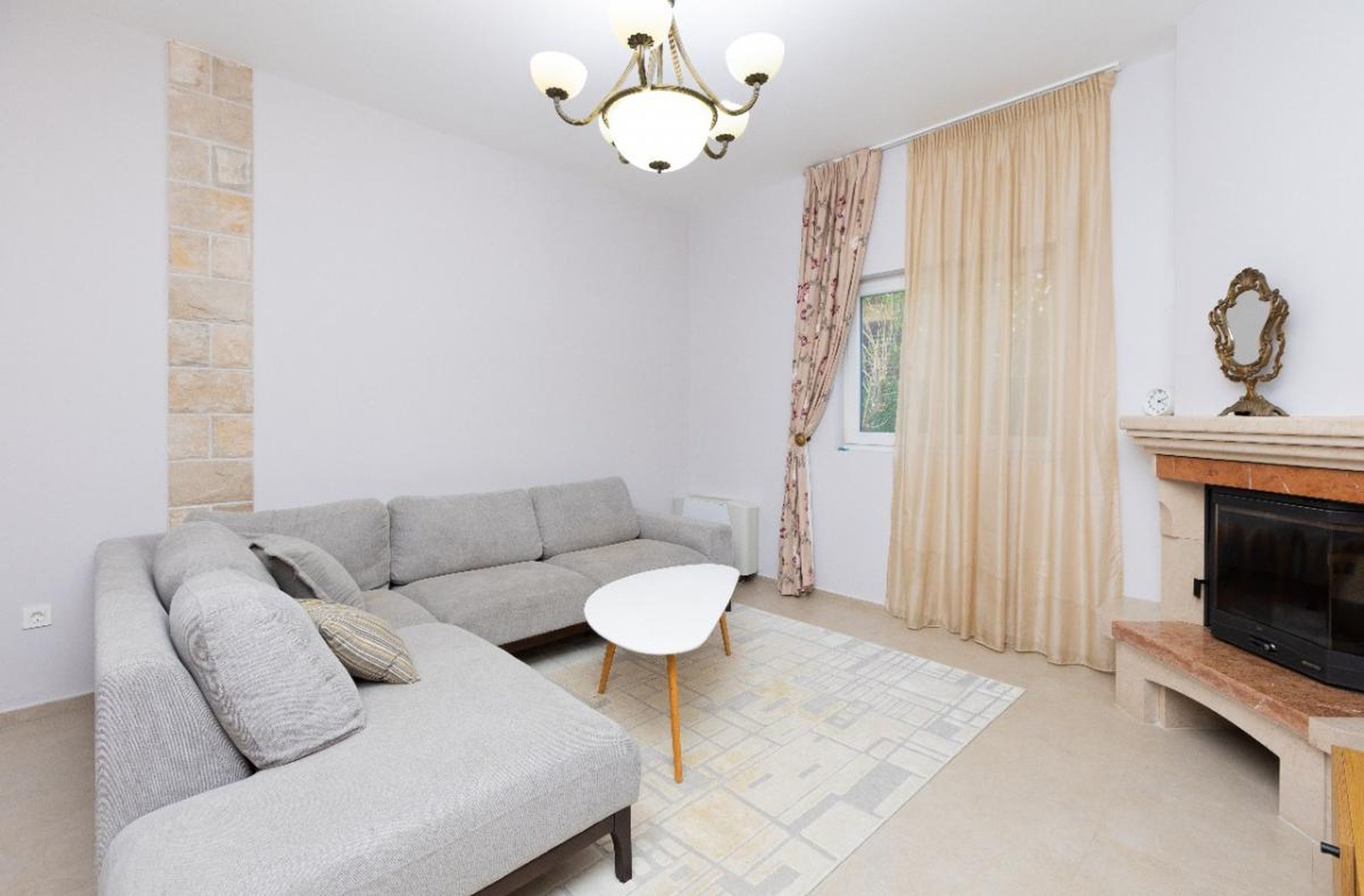 Salon dans une maison à Tivat, Monténégro : canapé d’angle gris, cheminée avec parement en pierre et intérieur clair.