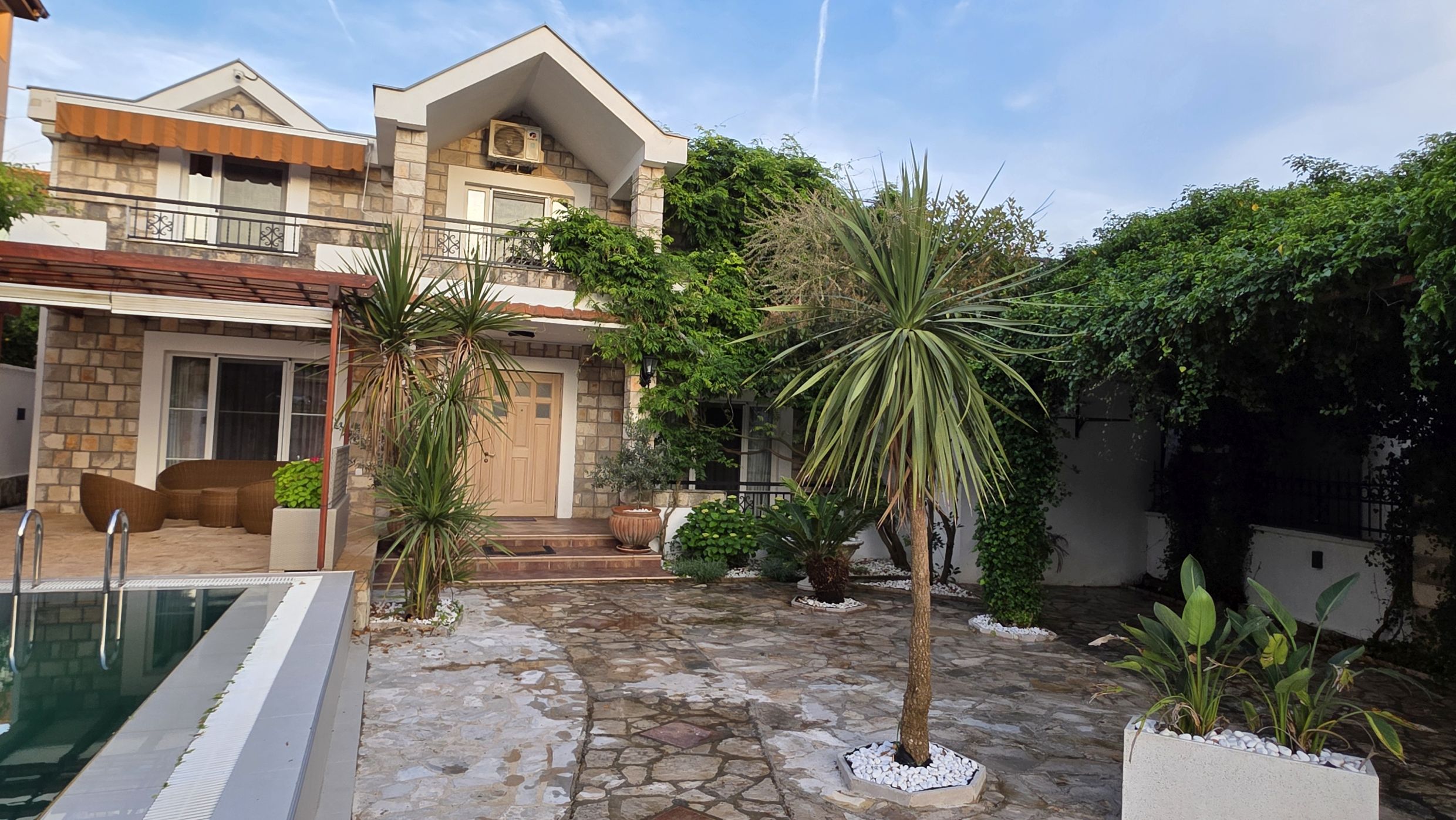 Maison à deux étages à Tivat, Monténégro : façade en pierre, terrasse avec piscine, palmiers et jardin bien entretenu.