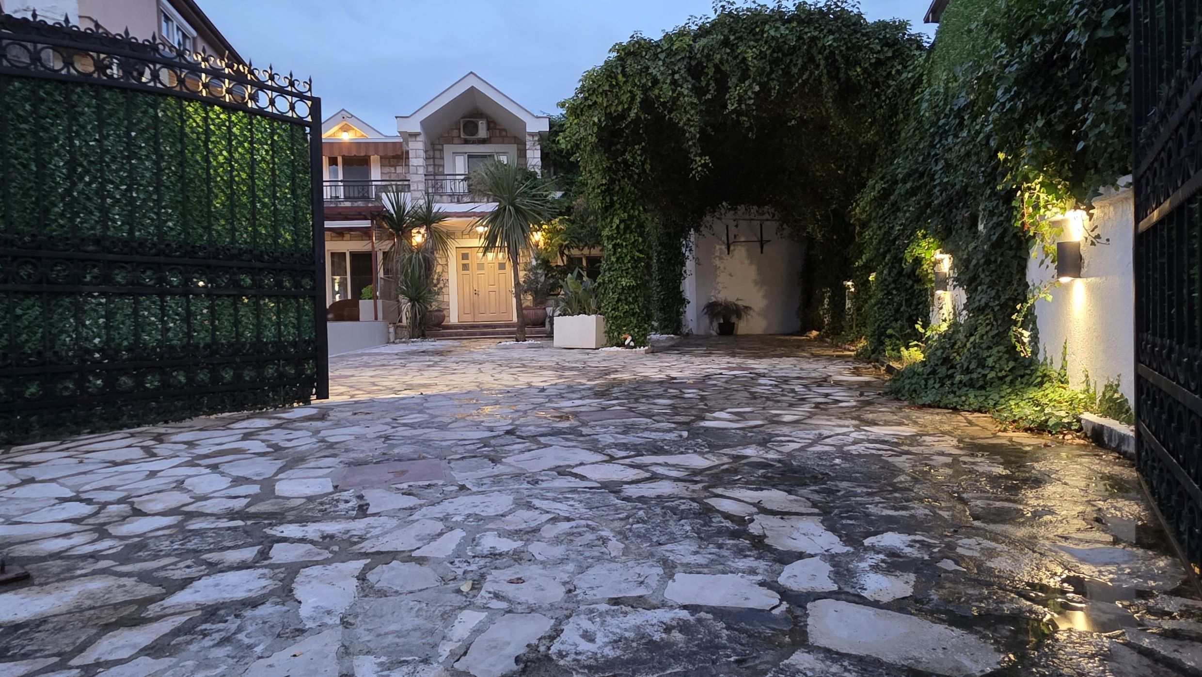 Cour d’une maison privée à Tivat, Monténégro : aire pavée en pierre, portail en fer forgé et arche verte de plantes grimpantes.