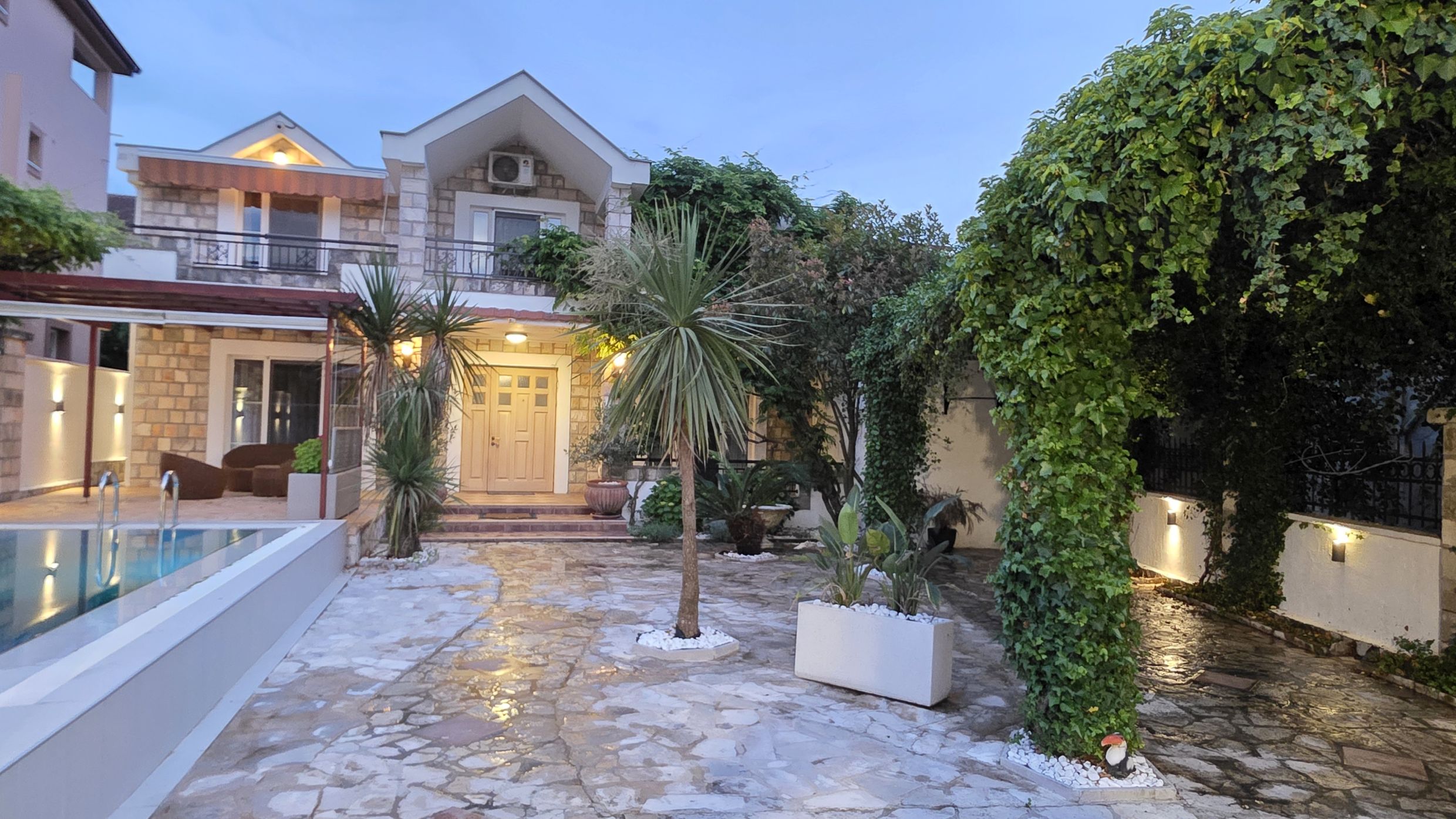 Villa de deux étages à Tivat, Monténégro : façade en pierre, piscine, jardin bien entretenu avec des palmiers et terrasse conviviale.
