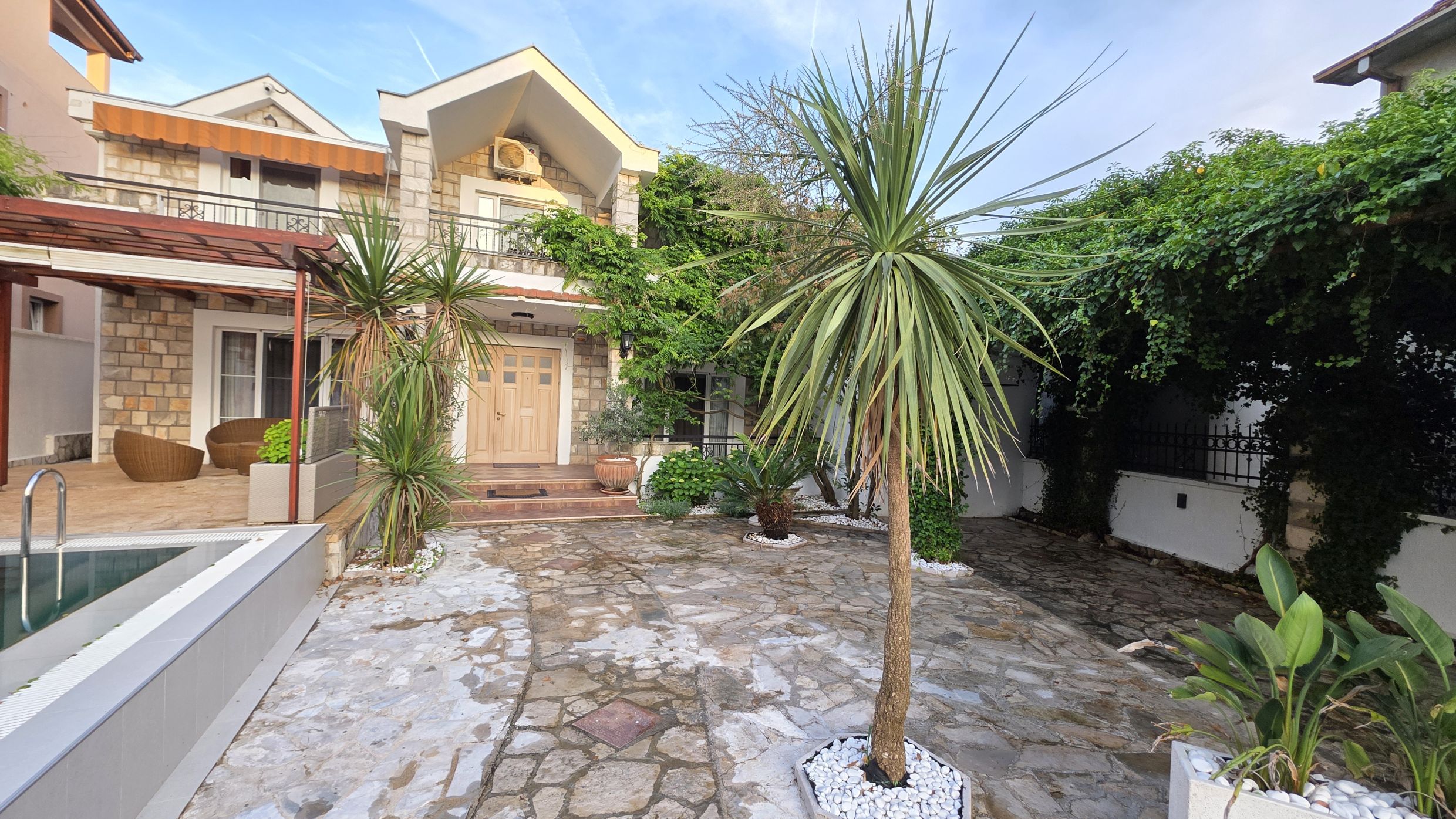 Maison à deux étages à Tivat, Monténégro : façade en pierre, terrasse avec mobilier en rotin, piscine et jardin bien entretenu.