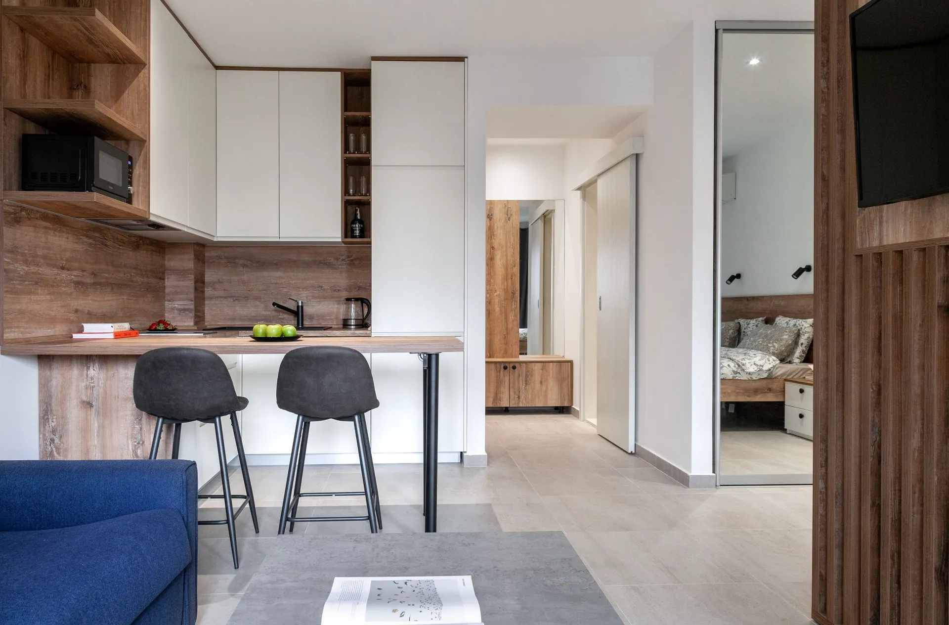 Modern kitchen with a wooden countertop, white cabinets, and black bar stools. A bedroom is visible in the mirror reflection.