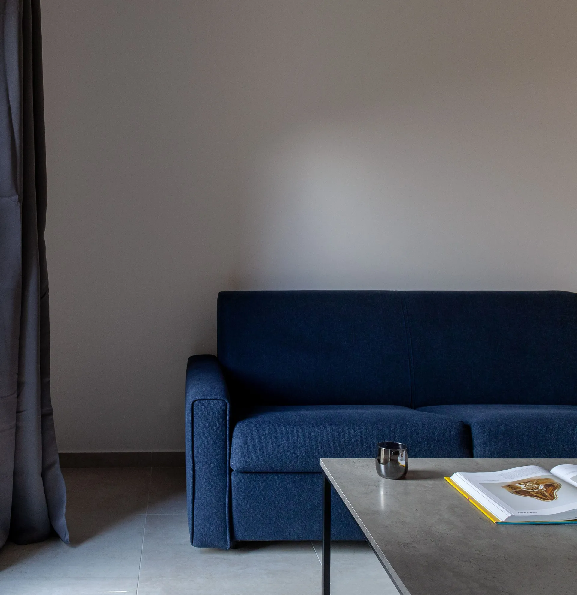 A dark blue sofa against a grey wall, with an open book and metal cup on the table, grey curtains on the left.