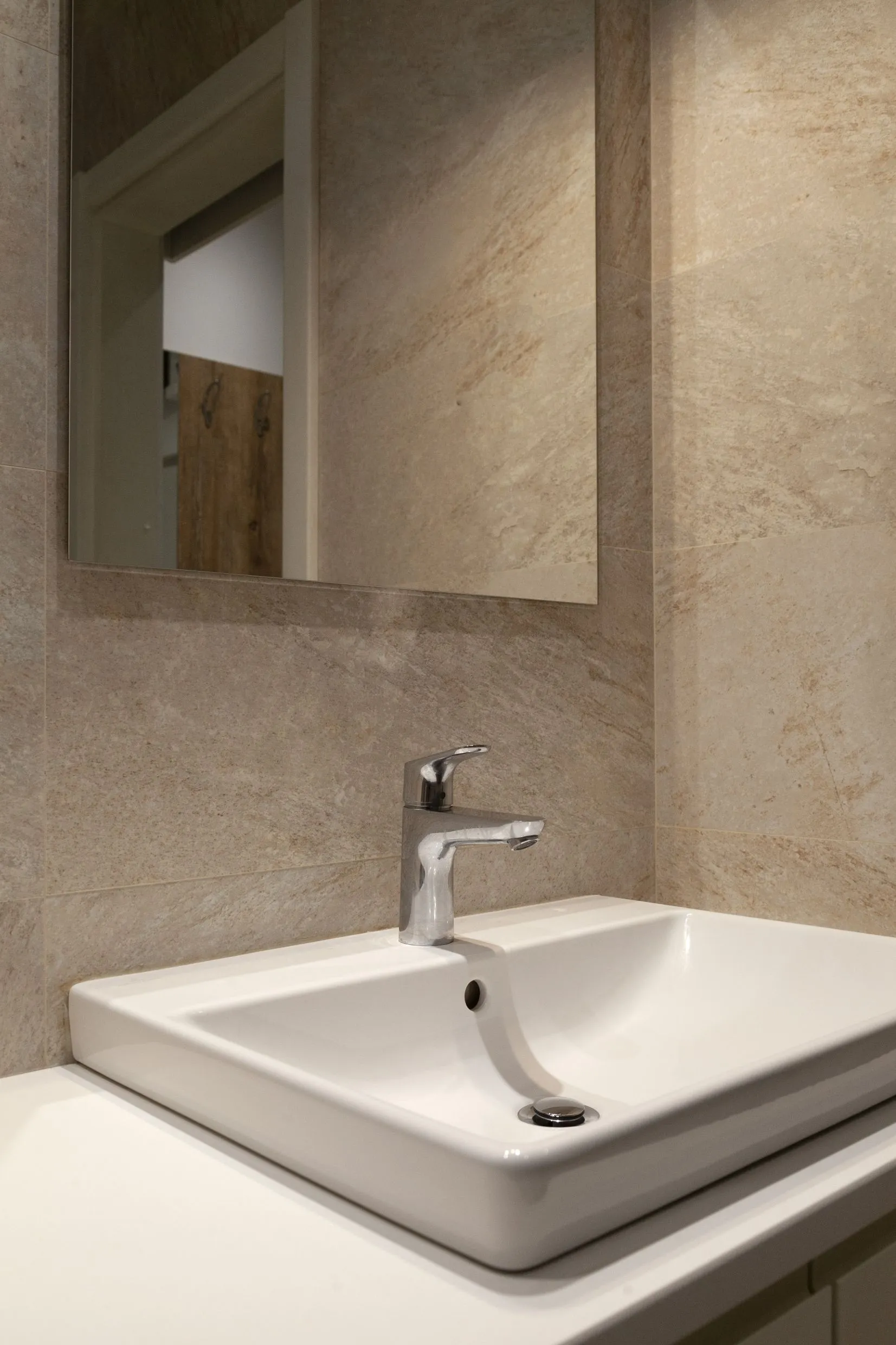White rectangular vessel sink with a chrome faucet set against beige stone tiles and a wall mirror.