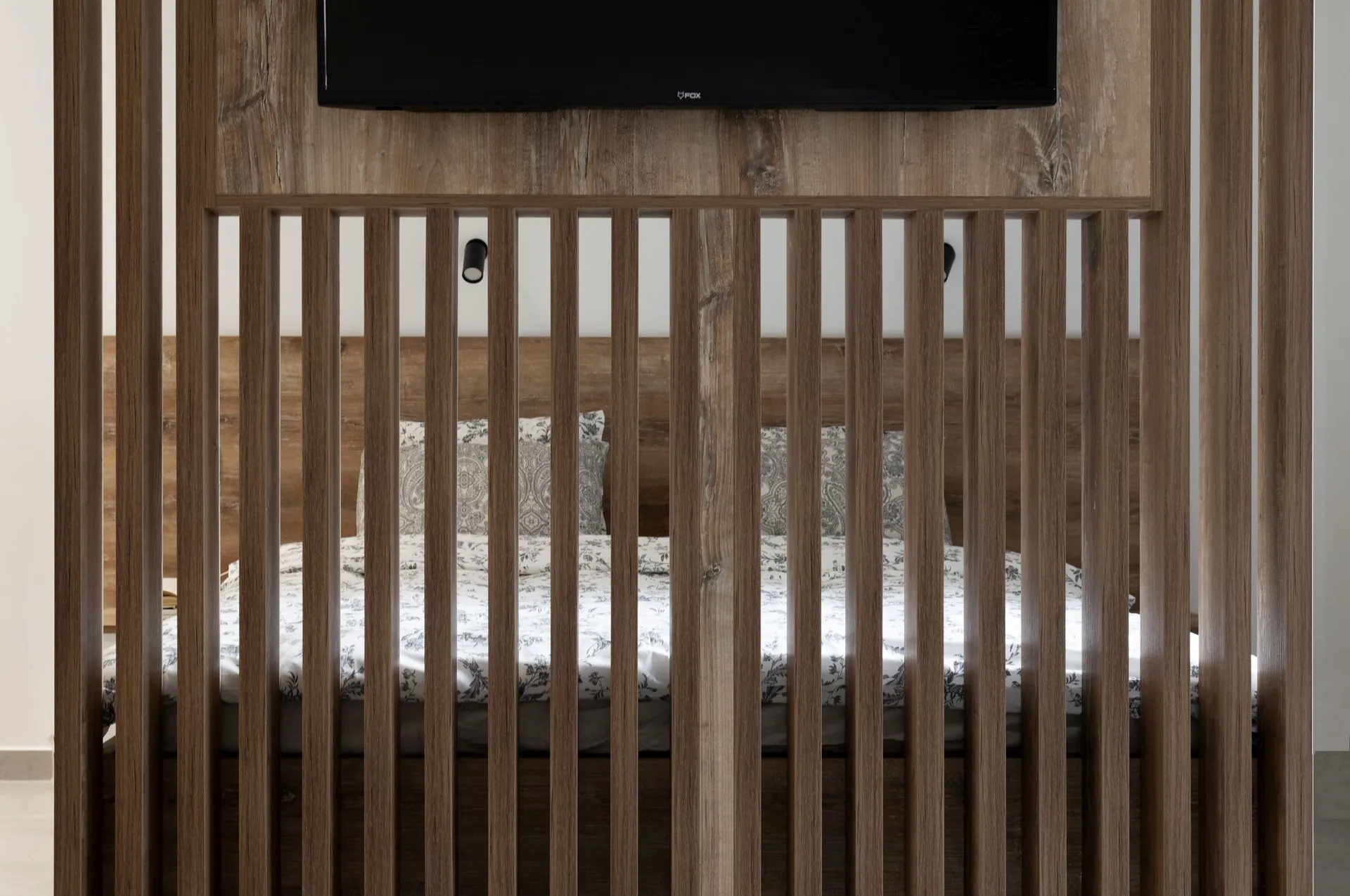 Wooden slat partition in front of a bed with floral bedding and a television mounted above.