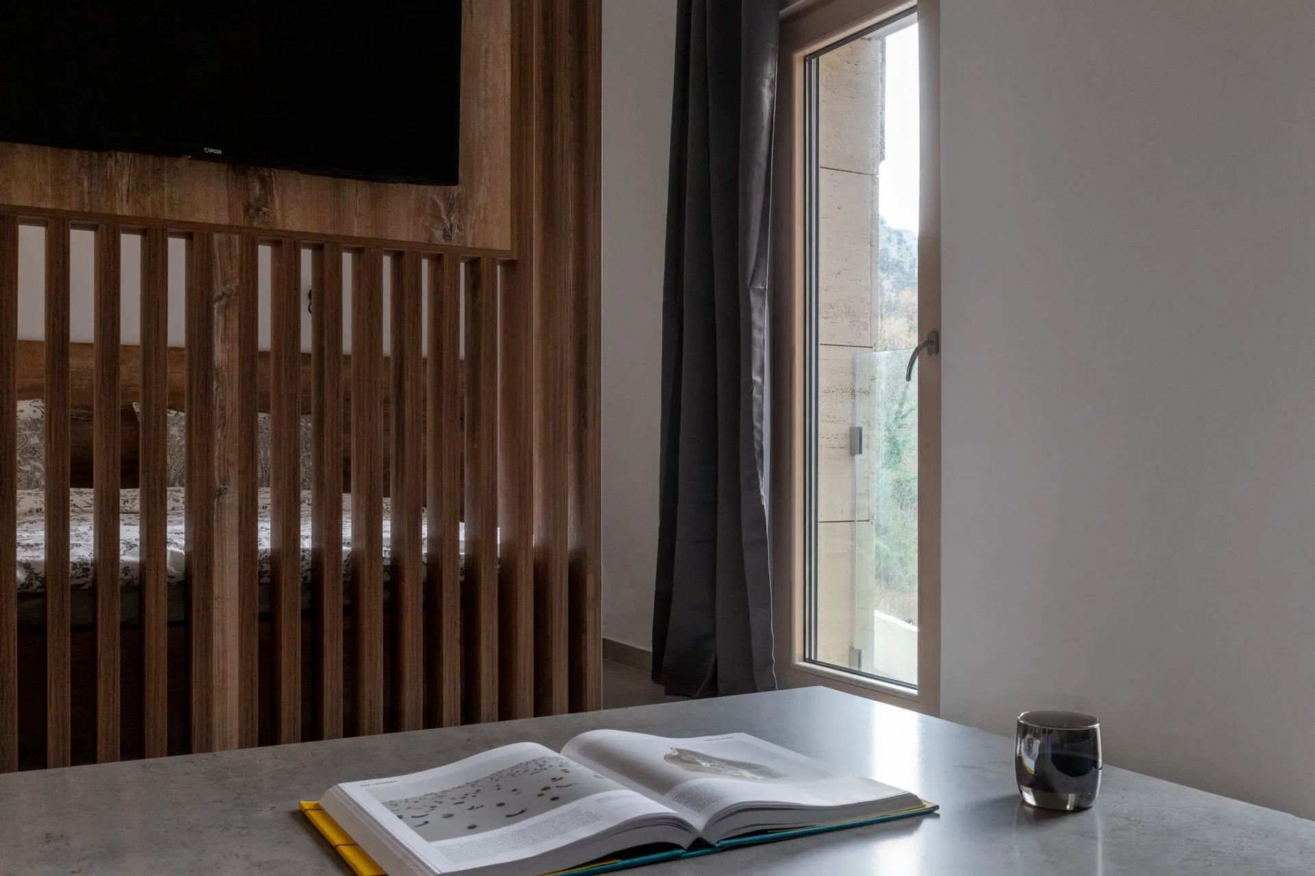 Modern bedroom with a wooden slat partition revealing the bed and TV. An open book and metal mug sit on a concrete desk next to a tall window.