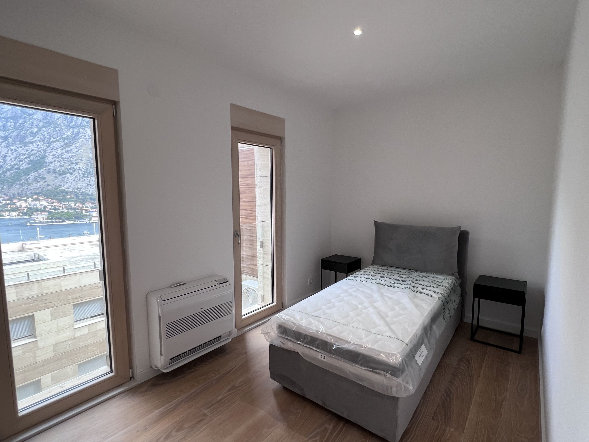 Schlafzimmer in einer Wohnung in Kotor, Montenegro: Bett mit Matratze, schwarze Nachttische, Panoramablick auf die Bucht von Kotor und die Berge.