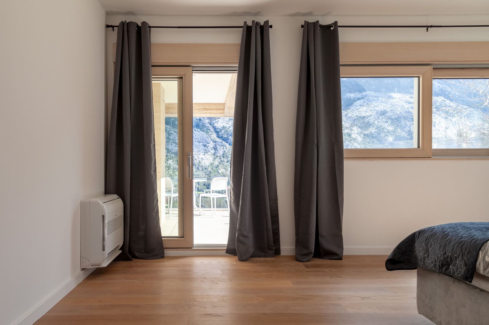 Schlafzimmer in einer Wohnung in Kotor, Montenegro: Panoramafenster, Zugang zur Terrasse mit Bergblick, Parkett und graue Vorhänge.