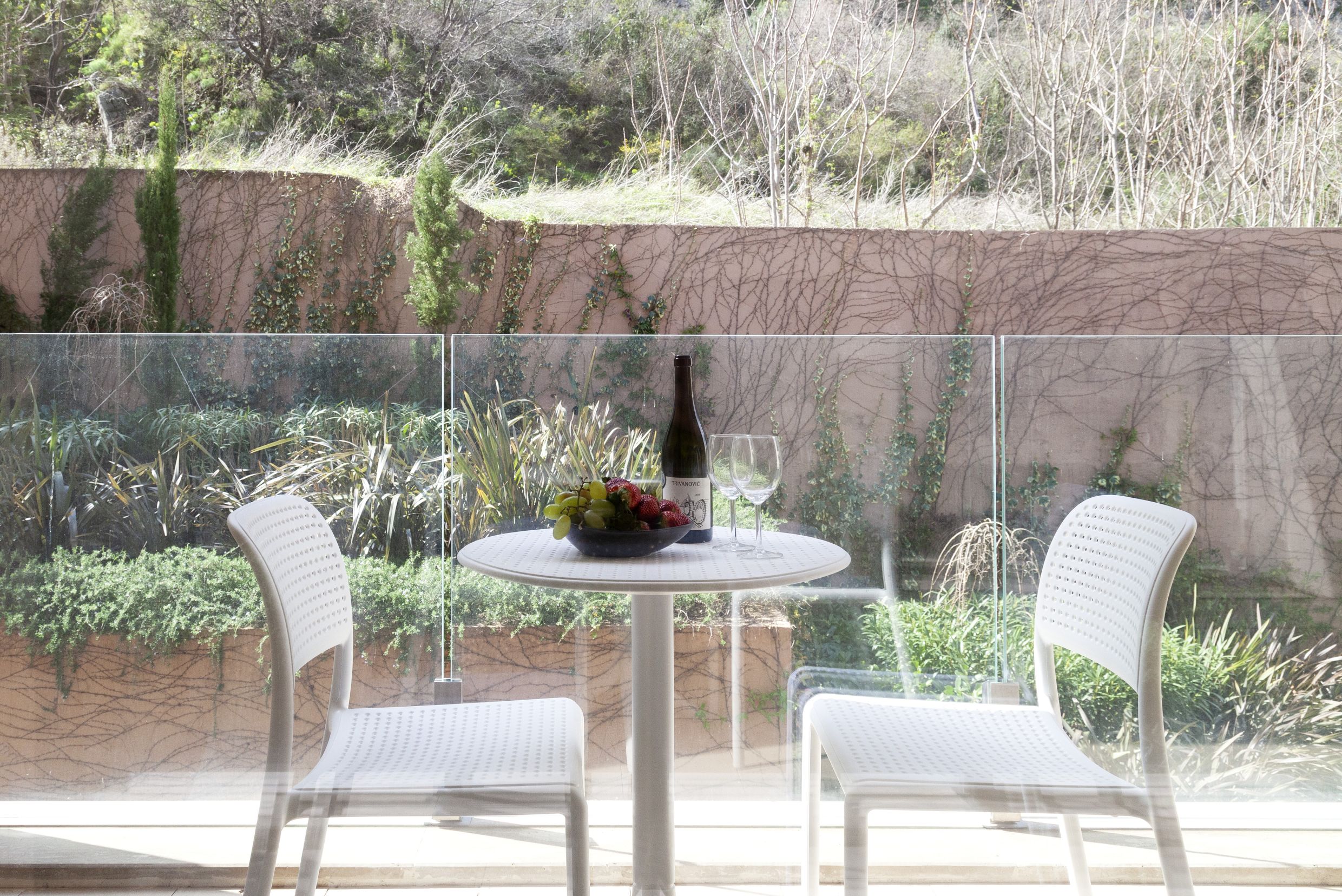 Balkon mit weißen Möbeln und Blick auf den Garten in einer Wohnung in Kotor, Montenegro: Tisch mit Wein und Früchten hinter Glas.