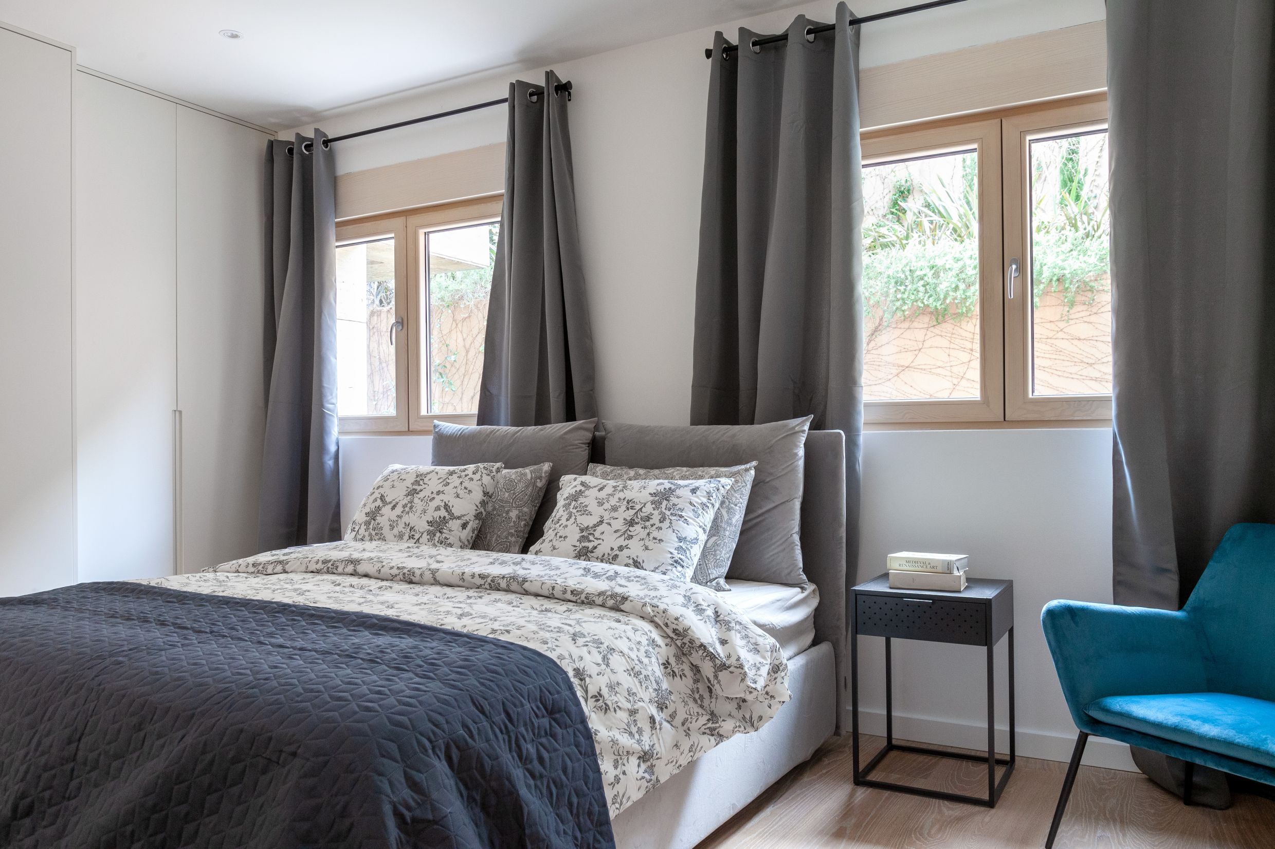 Modernes Schlafzimmer in einer Wohnung in Kotor, Montenegro: Doppelbett, blauer Sessel und Blick auf den Garten aus den Fenstern.