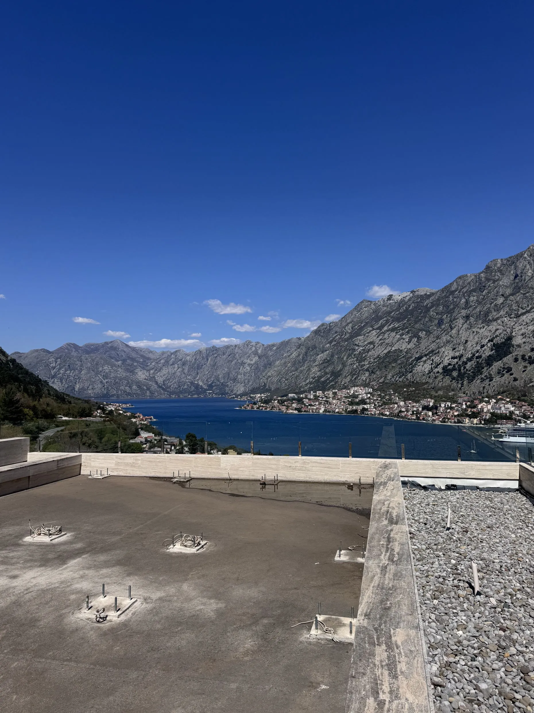Open terrace with a view of a mountain bay and town. Foreground shows concrete surface with metal fixtures and a glass safety barrier.