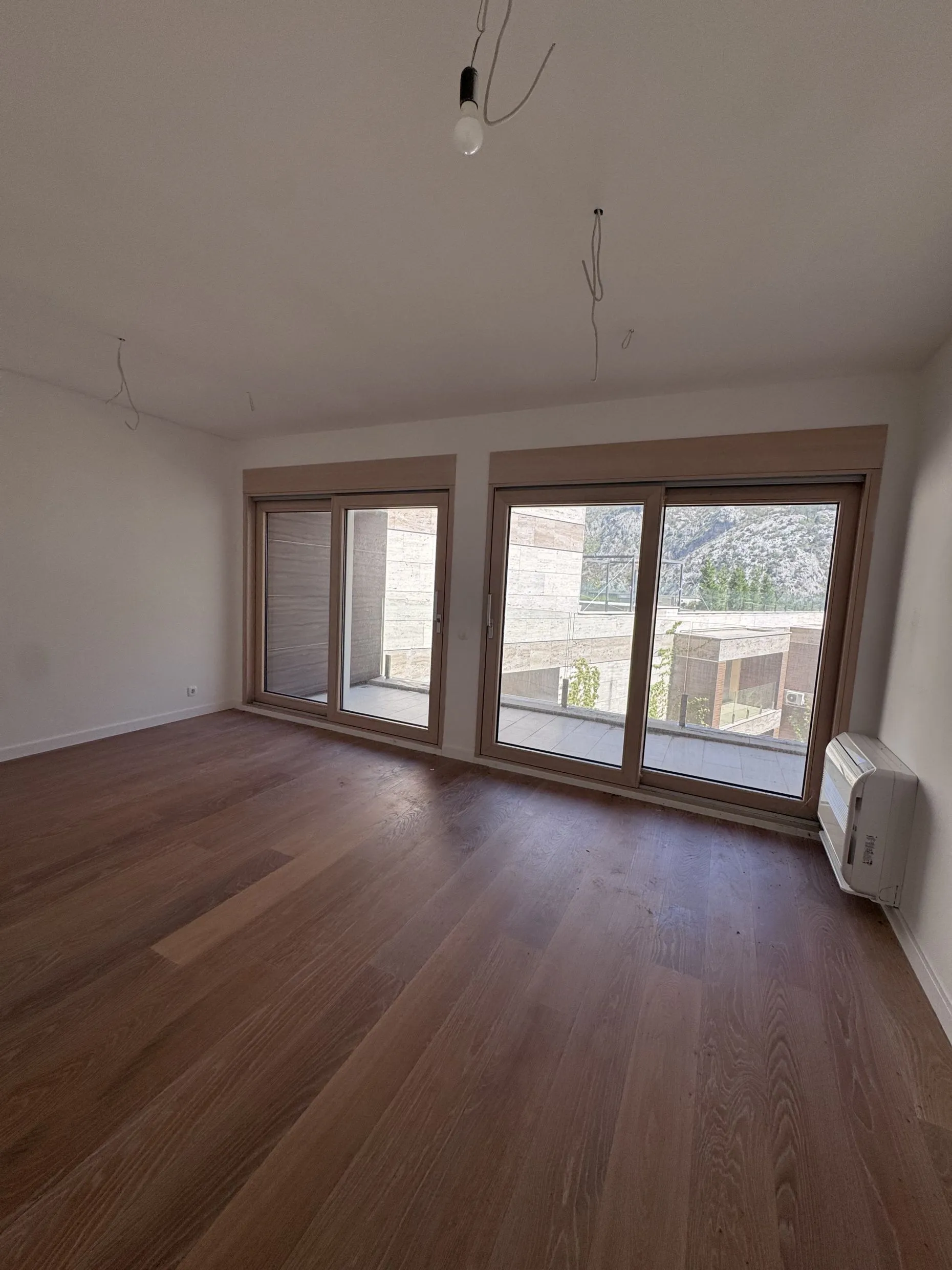 Empty room with wooden flooring and large windows. Exposed ceiling wires and a wall-mounted air conditioner are visible.